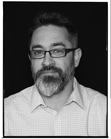 Black and white portrait of a man with glasses, short hair, beard, and mustache, wearing a checkered shirt.