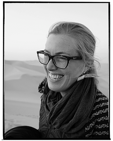 Black and white photo of a smiling woman with glasses outdoors in desert landscape.