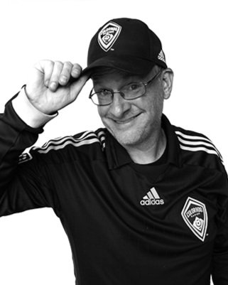 A man wearing glasses, a black Adidas jacket, a black cap with a badge, and a badge on his chest, smiling and tipping his cap.
