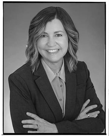 Black and white professional headshot of a woman with shoulder-length wavy hair, wearing a blazer and a collared shirt, smiling with arms crossed.
