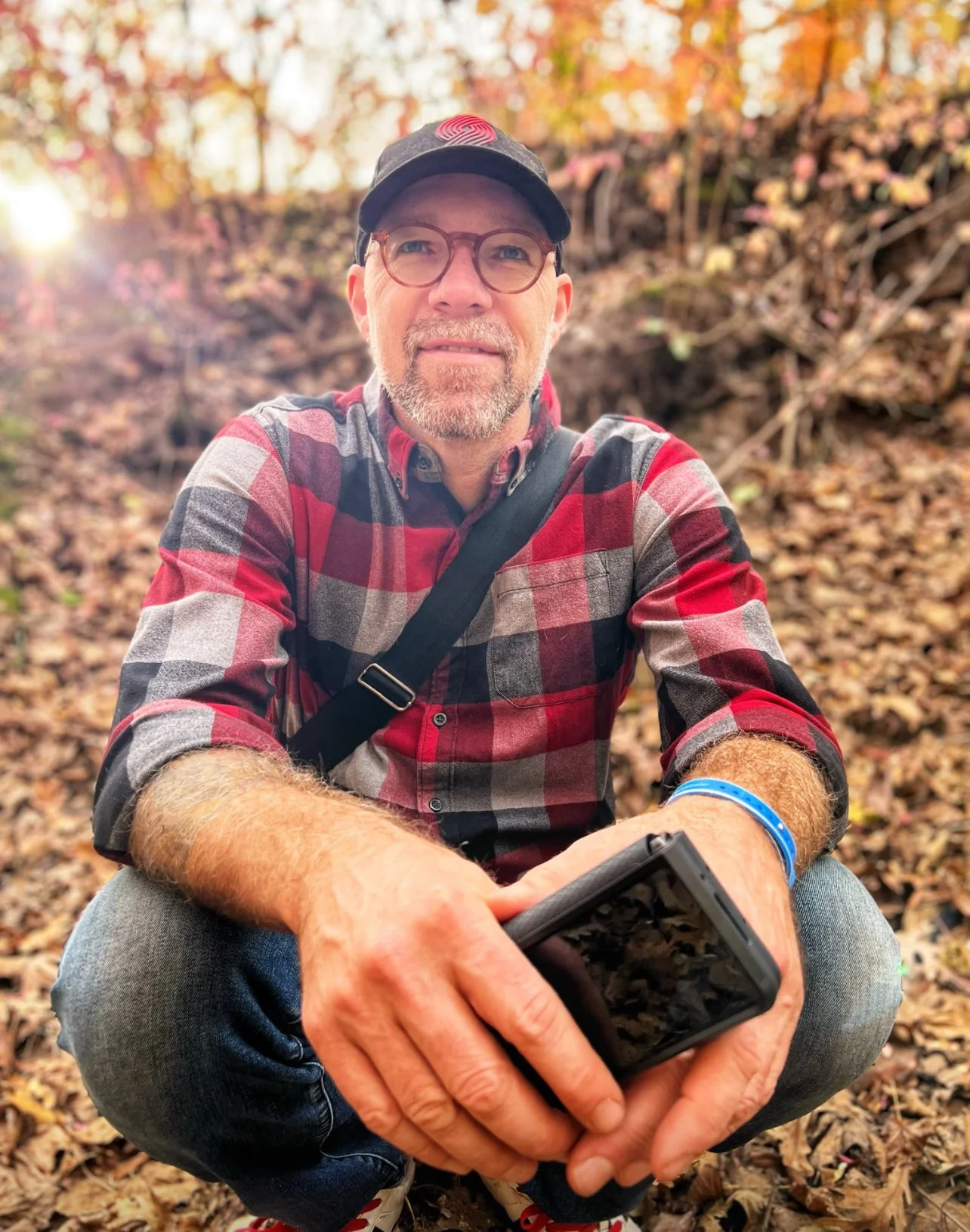 Man in plaid shirt and cap sitting in autumn foliage, holding a phone