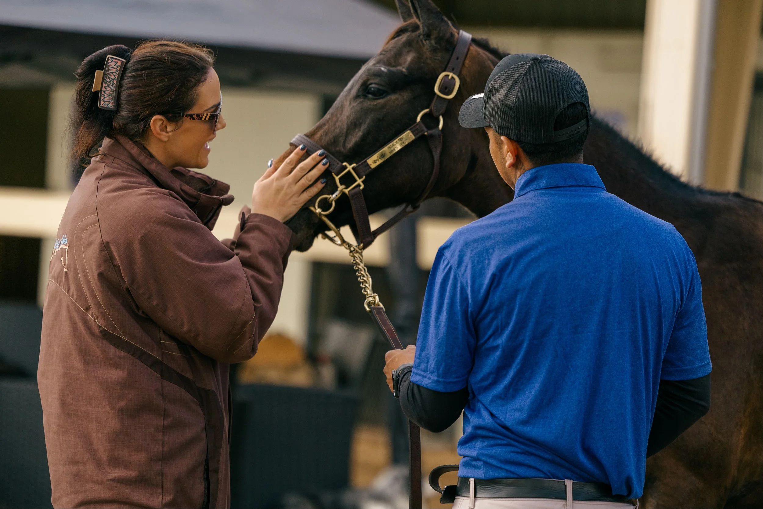 A woman petting a brown horse while a man in a blue shirt holds the horse's lead in an outdoor stable area.