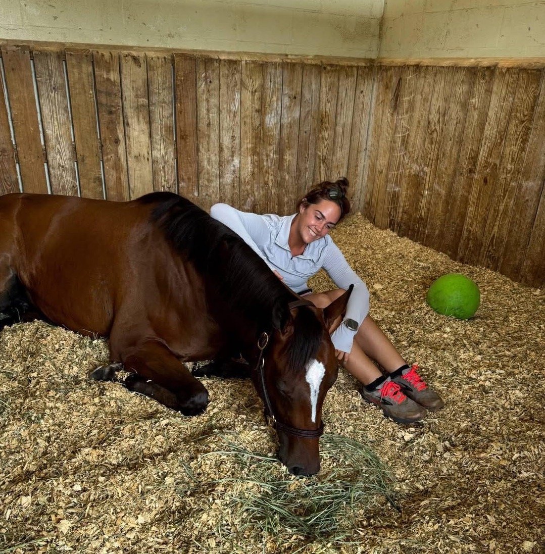 A woman in a light blue shirt and gray shorts sitting on hay in a wooden stall, smiling, with her arm around a resting brown horse with a white stripe on its face, near a green watermelon-shaped ball.