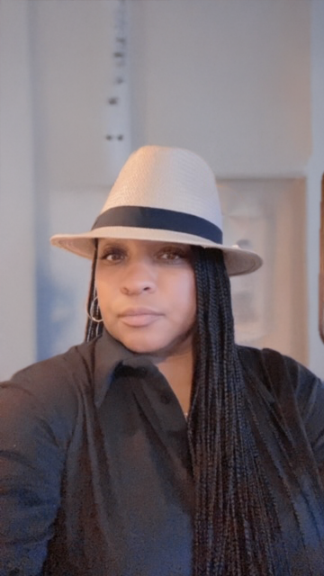Woman wearing a white fedora hat with a black band and a black button-up shirt, with long braided hair, indoors.
