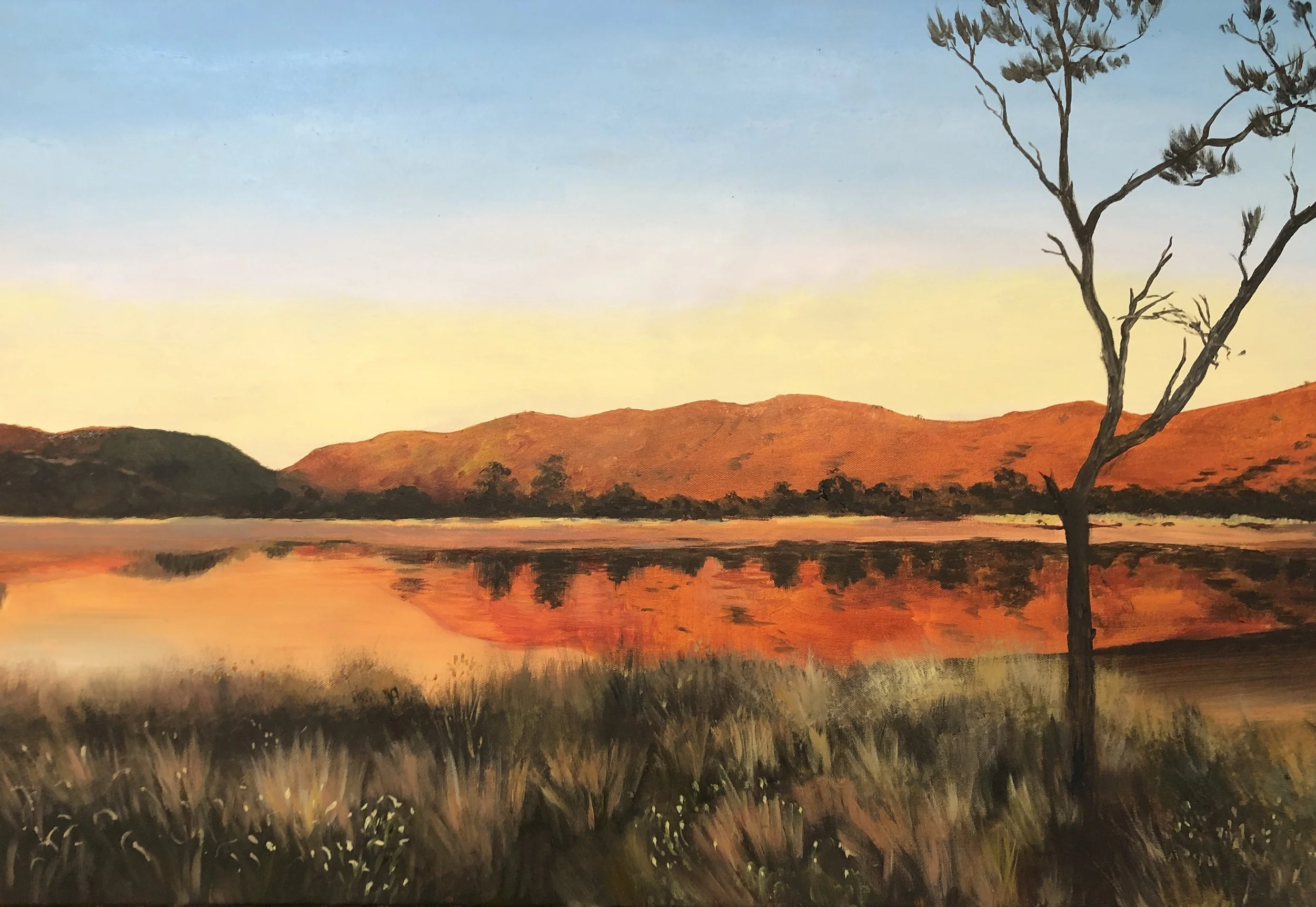 A landscape painting of a lake reflecting orange and red mountains during sunset, with a single leafless tree in the foreground and blue sky overhead.