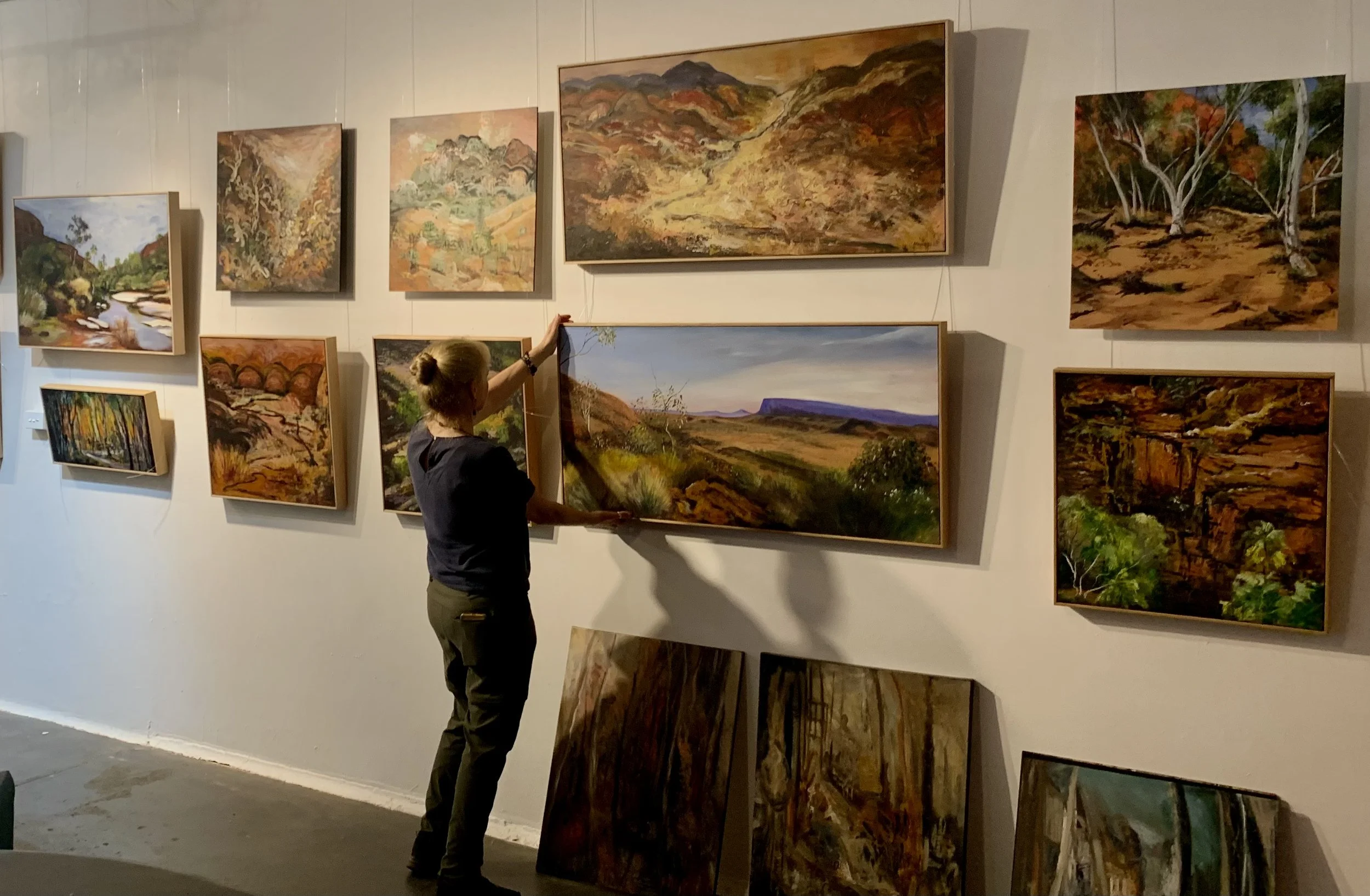 A woman arranging landscape paintings on a gallery wall, with various framed artworks depicting nature scenes and colorful landscapes