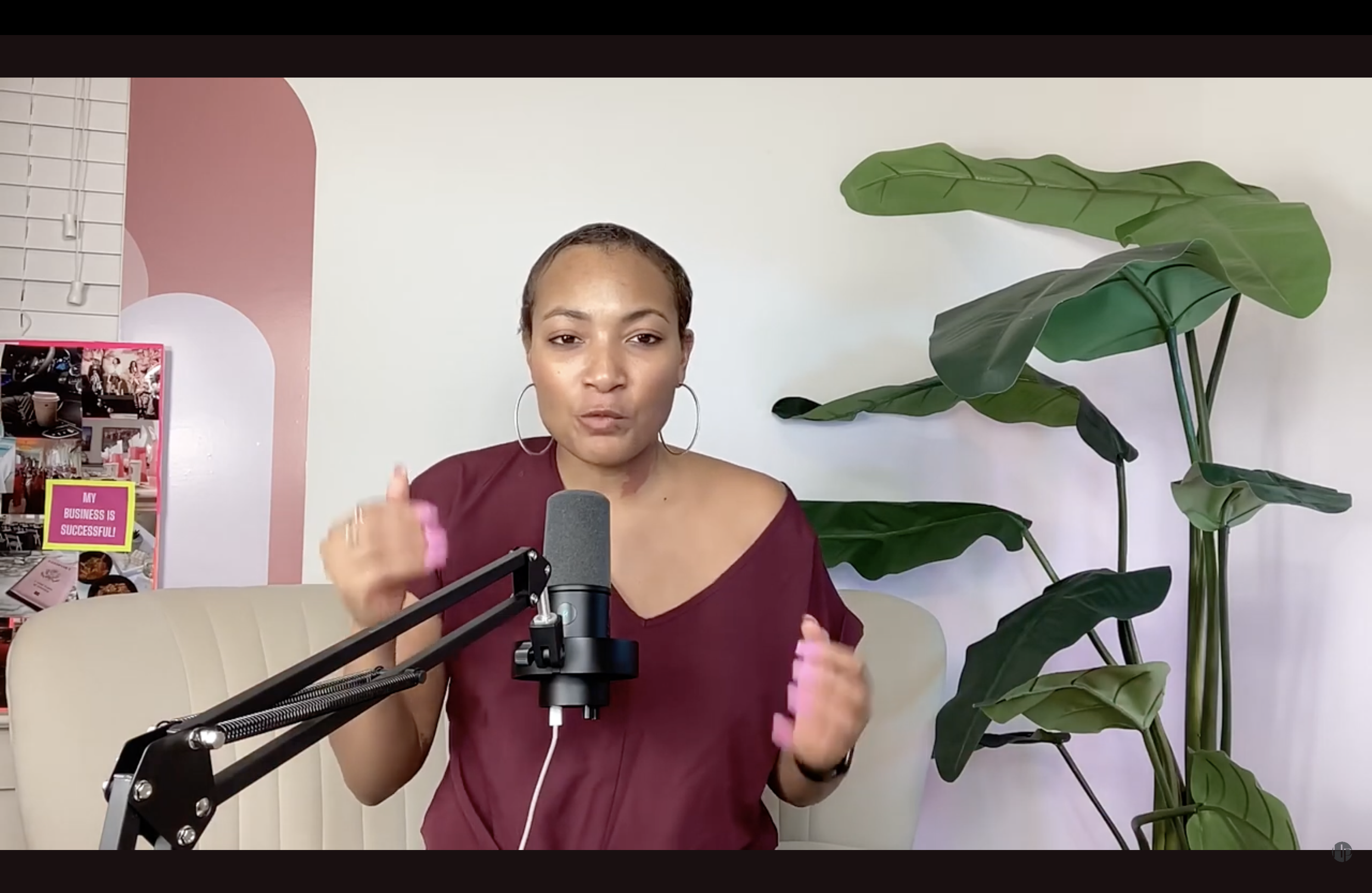 A woman with short hair, hoop earrings, and a maroon shirt is speaking into a microphone during a podcast recording. She wears a pink wristband on her right wrist. The background features a large green plant with broad leaves and a pink and white display stand with a sign that reads 'My Business is Successful!'