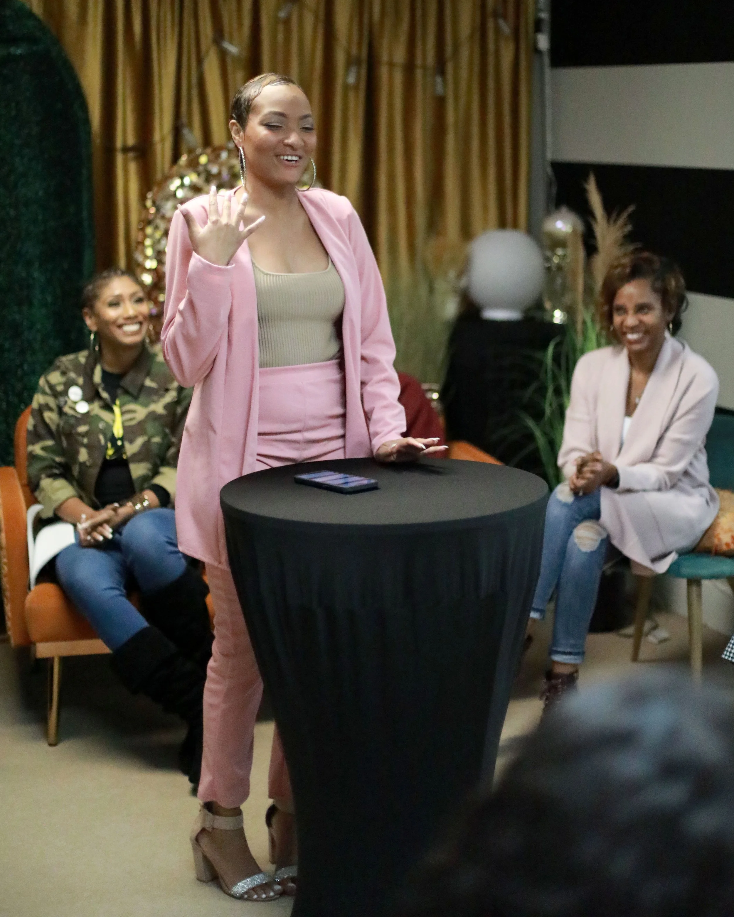 A woman in a pink blazer and matching pink pants is smiling and gesturing with her left hand during a social gathering. She has short hair and large hoop earrings. Behind her, two women are seated; one wearing a camouflage jacket and the other in a light pink blazer, both smiling. The setting appears to be indoors with decorative elements like gold curtains and plants.
