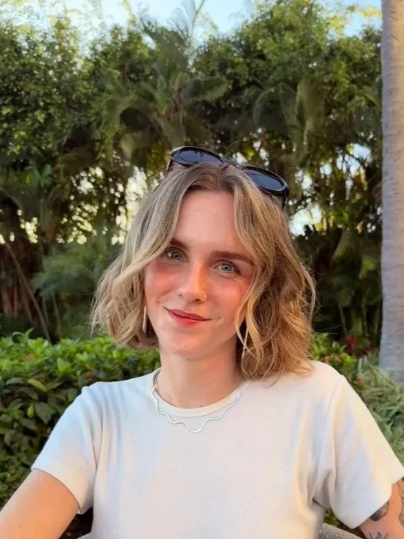 A woman with shoulder-length wavy blonde hair, wearing sunglasses on her head and a white t-shirt, sitting outdoors with green trees and plants in the background. She is a beautiful, educated preschool director.