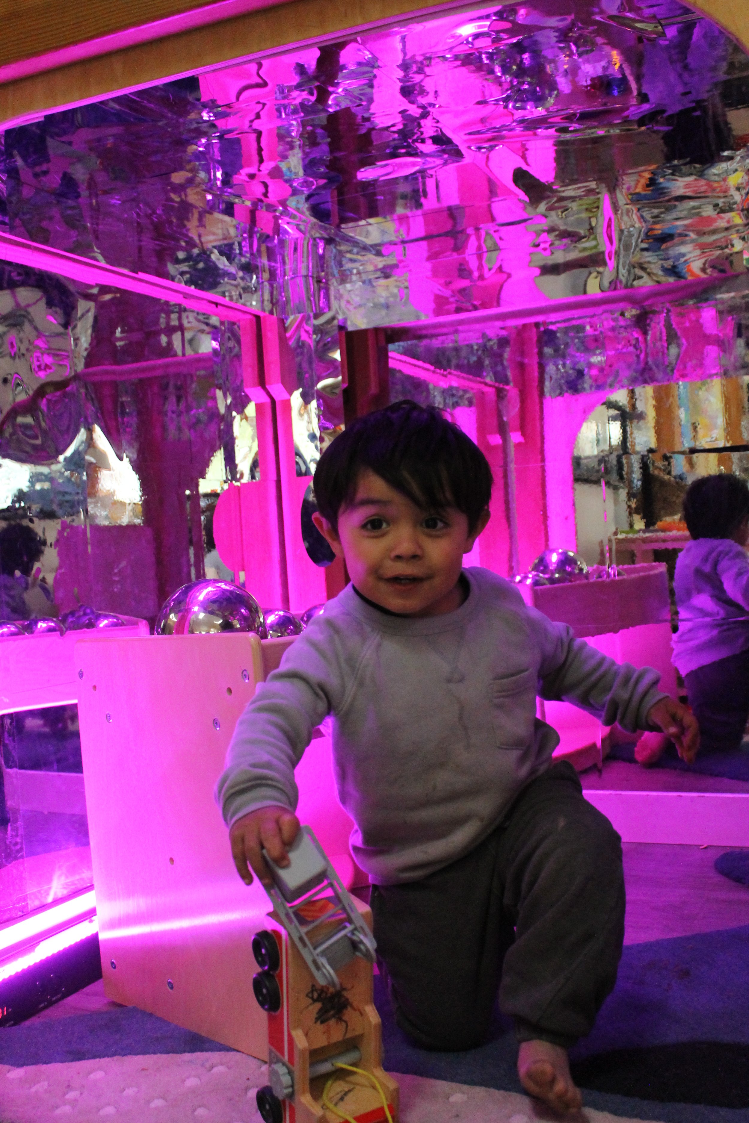 A young boy with dark hair sitting on the floor in front of a pink-lit mirror installation, holding a toy car, with a playful expression, and another child visible in the background.