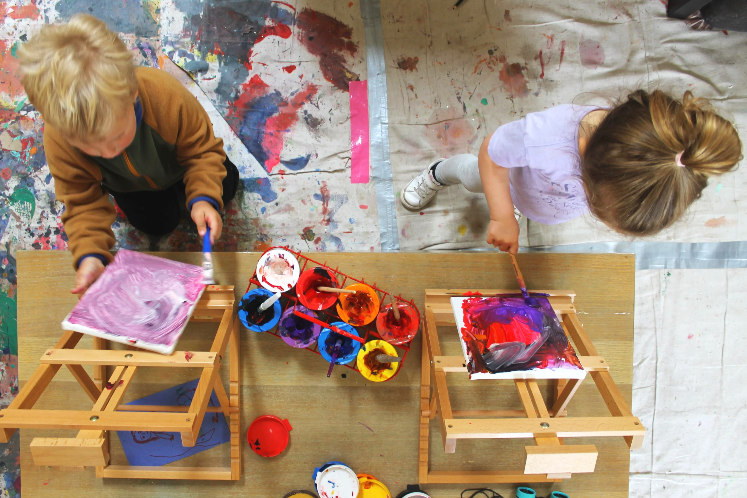 Two children painting on small canvases at an art station with paint cups and brushes, on a splattered table cover.