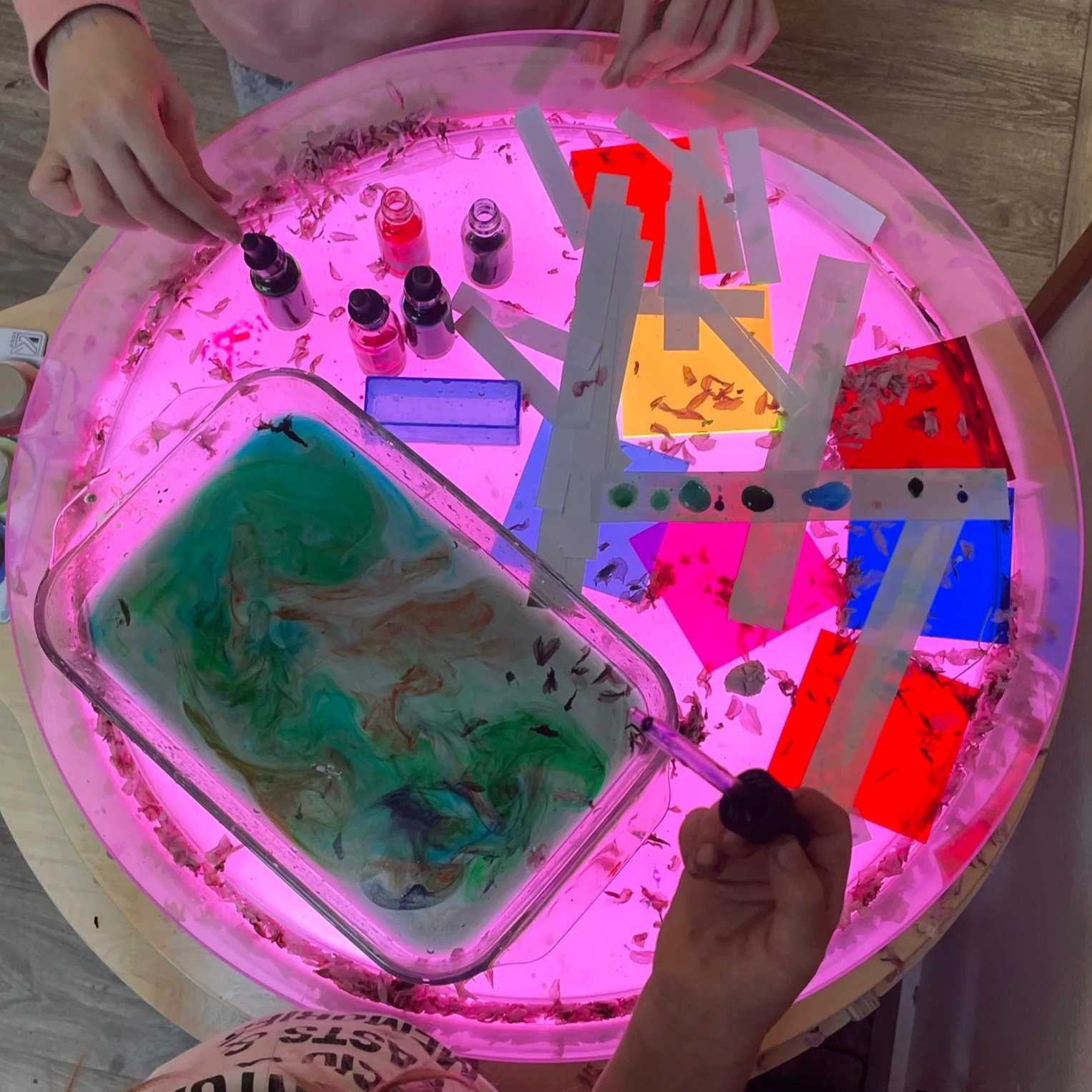 Child playing with oil and water in a sensory light table with a pink-lit circular surface, with bottles of colored liquid and various colored paper pieces.