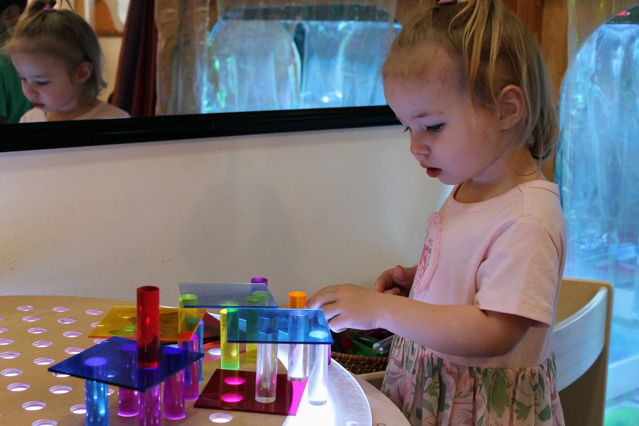 A young girl with blonde hair wearing a pink dress, concentrating on a colorful light table with various acrylic pieces. There is a mirror behind her reflecting her face and part of her surroundings.