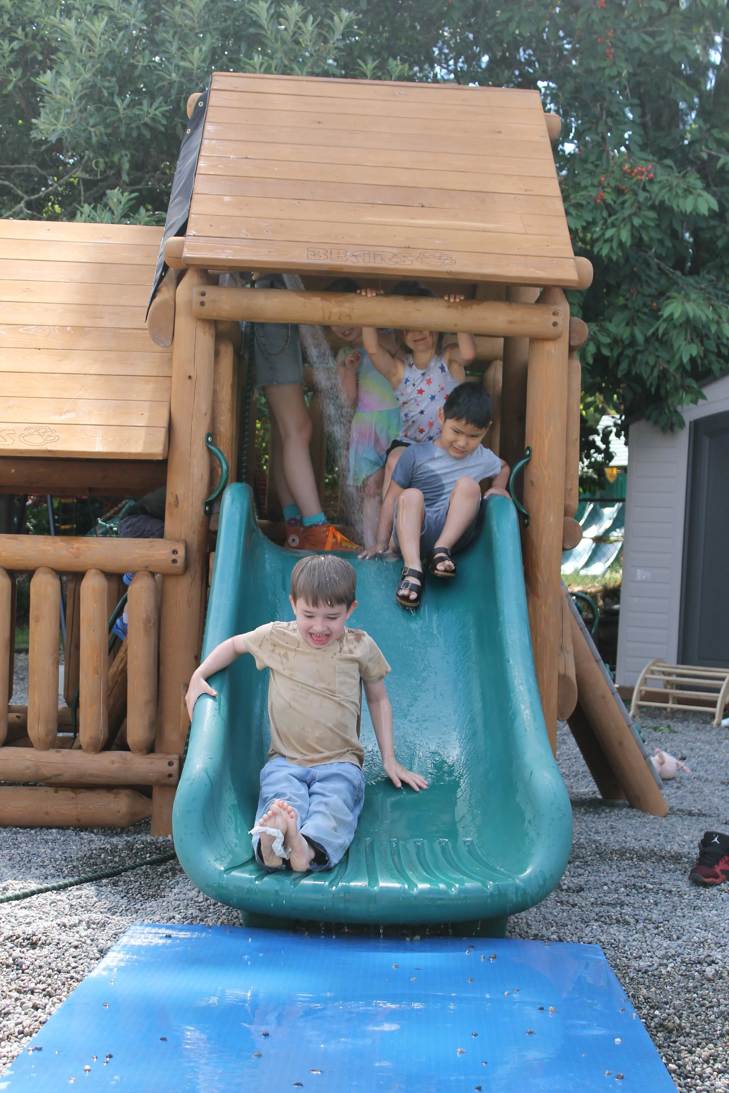 Children playing on a wooden playground slide outdoors, with some kids coming down the slide and others waiting at the top, trees in the background, and a blue cushioned mat at the bottom.