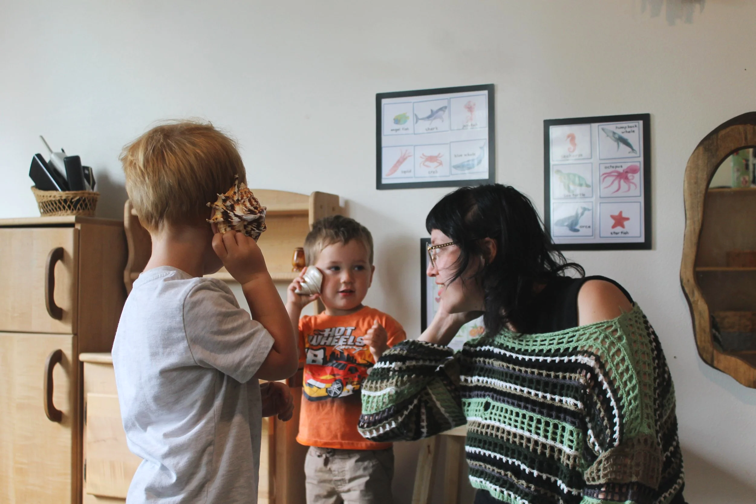 Three young children and an adult woman engaging in an activity indoors. One child is holding a seashell to his ear, the woman is smiling, and another child looks on. The background has framed pictures of sea animals on the wall.