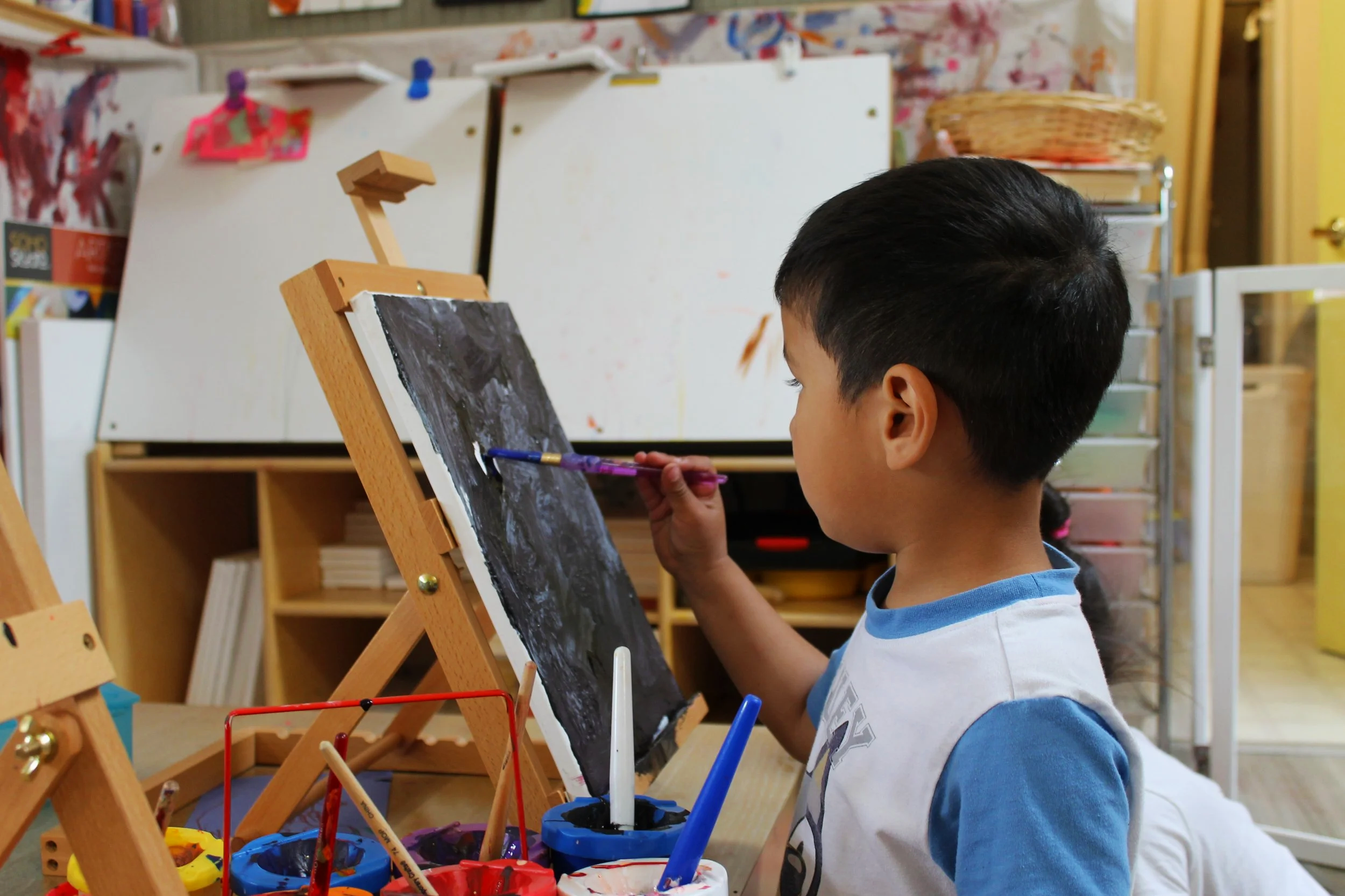 A young boy with dark hair and a blue and white shirt painting on a small chalkboard easel in a classroom or art studio. Paint cups and brushes are in front of him, and colorful supplies and storage shelves are in the background.