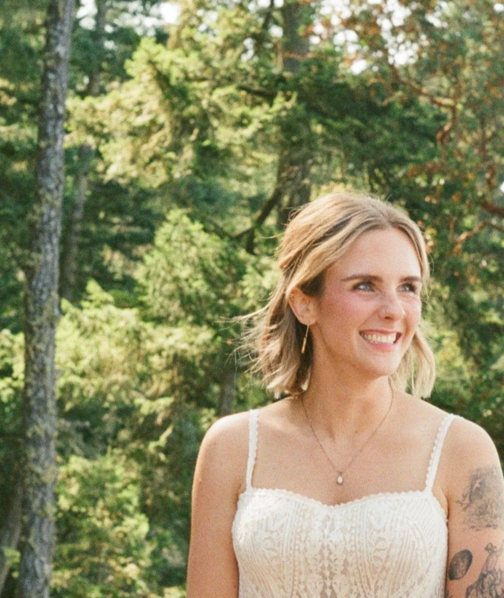 A woman with shoulder-length blonde hair smiling outdoors in a wooded area, wearing a white sleeveless dress and jewelry.