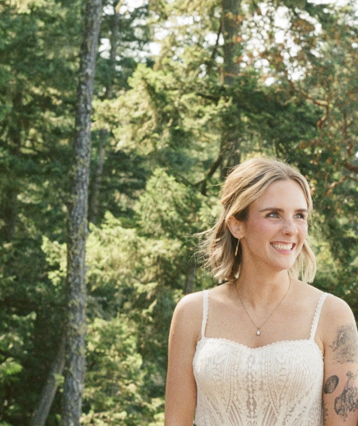 A young woman with short, wavy hair, wearing a white dress, standing outdoors in a wooded area with green leaves and tall trees in the background, smiling at the camera.