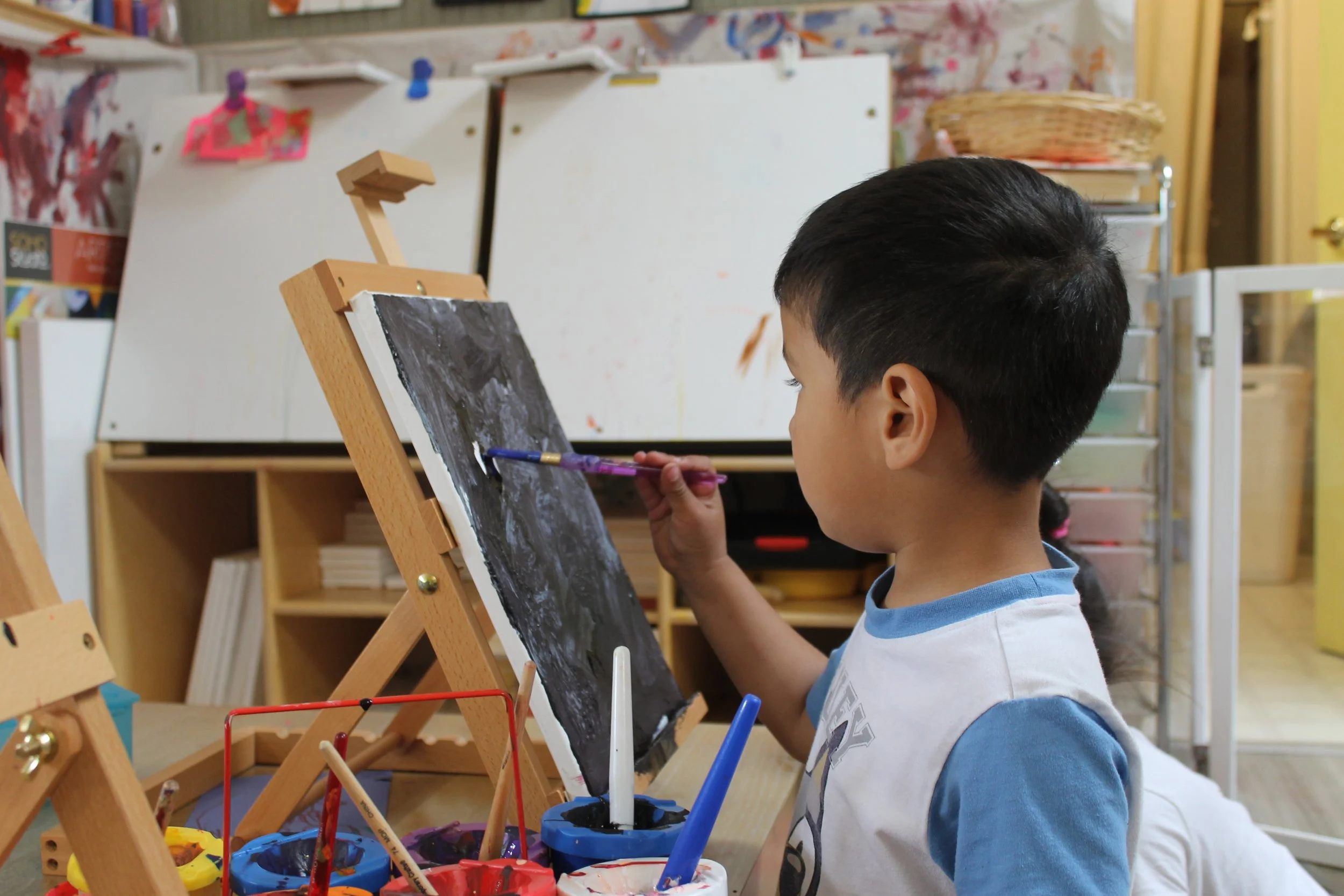 A young boy painting on a black canvas set up on a small wooden easel in an art classroom, with various paint containers and brushes in front of him.