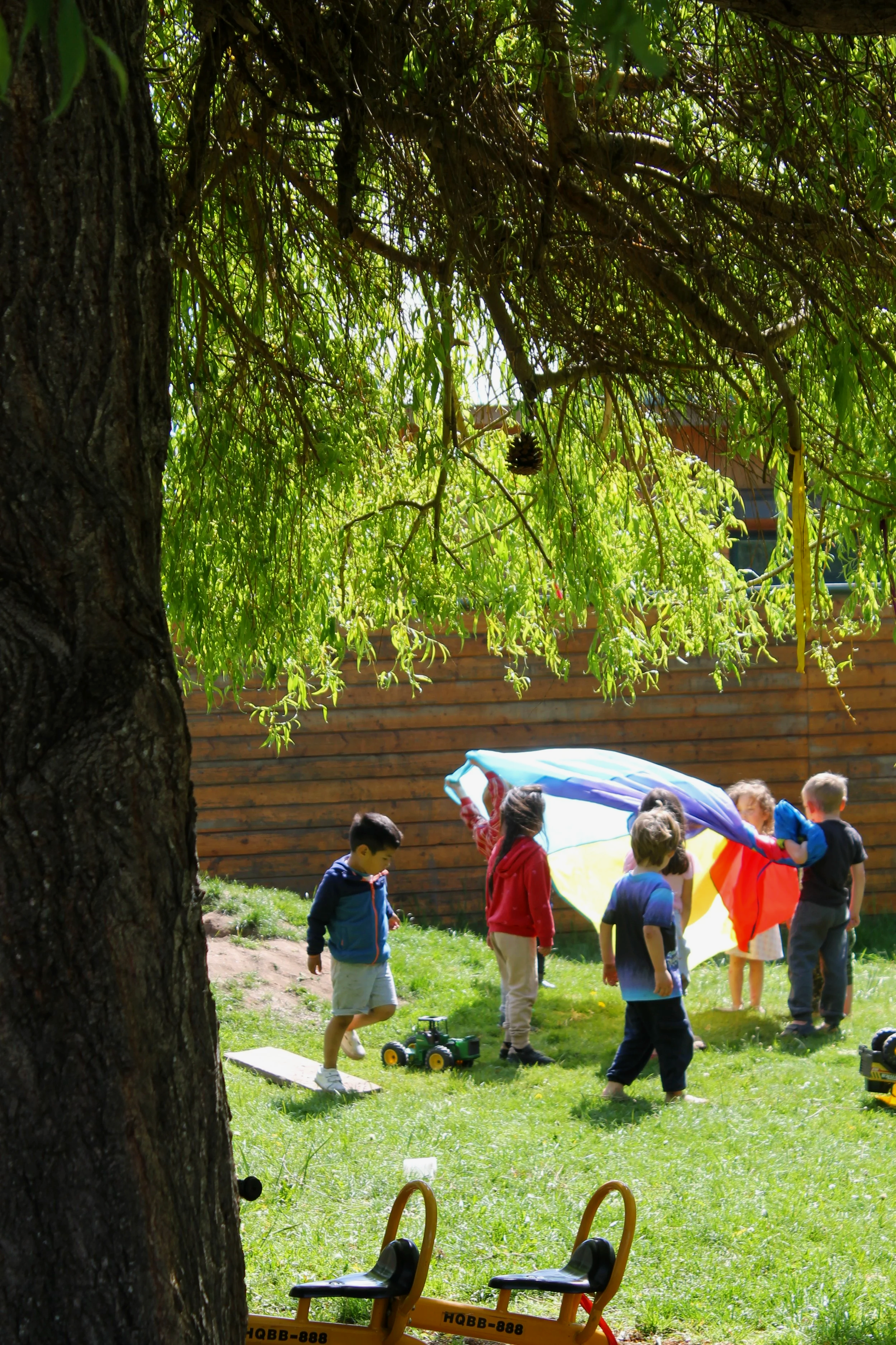 Children playing outdoors with a colorful parachute in a backyard, surrounded by trees and a wooden fence.
