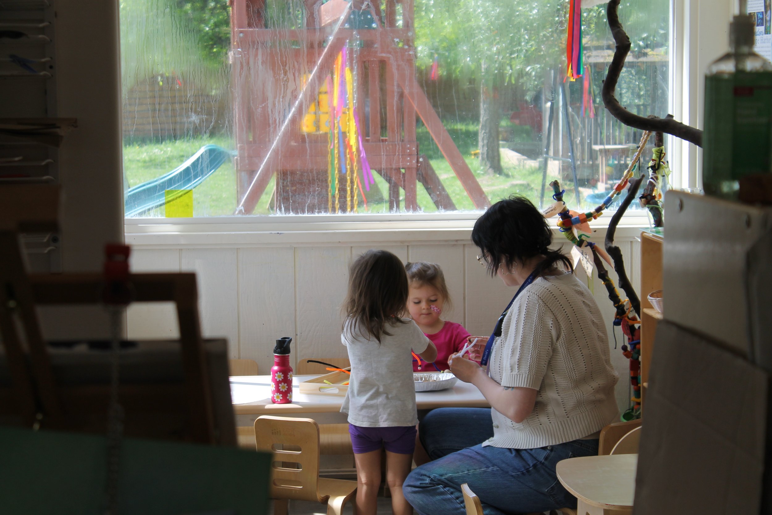 Children and a woman interacting at a table inside a classroom, with a window showing an outdoor playground and trees.