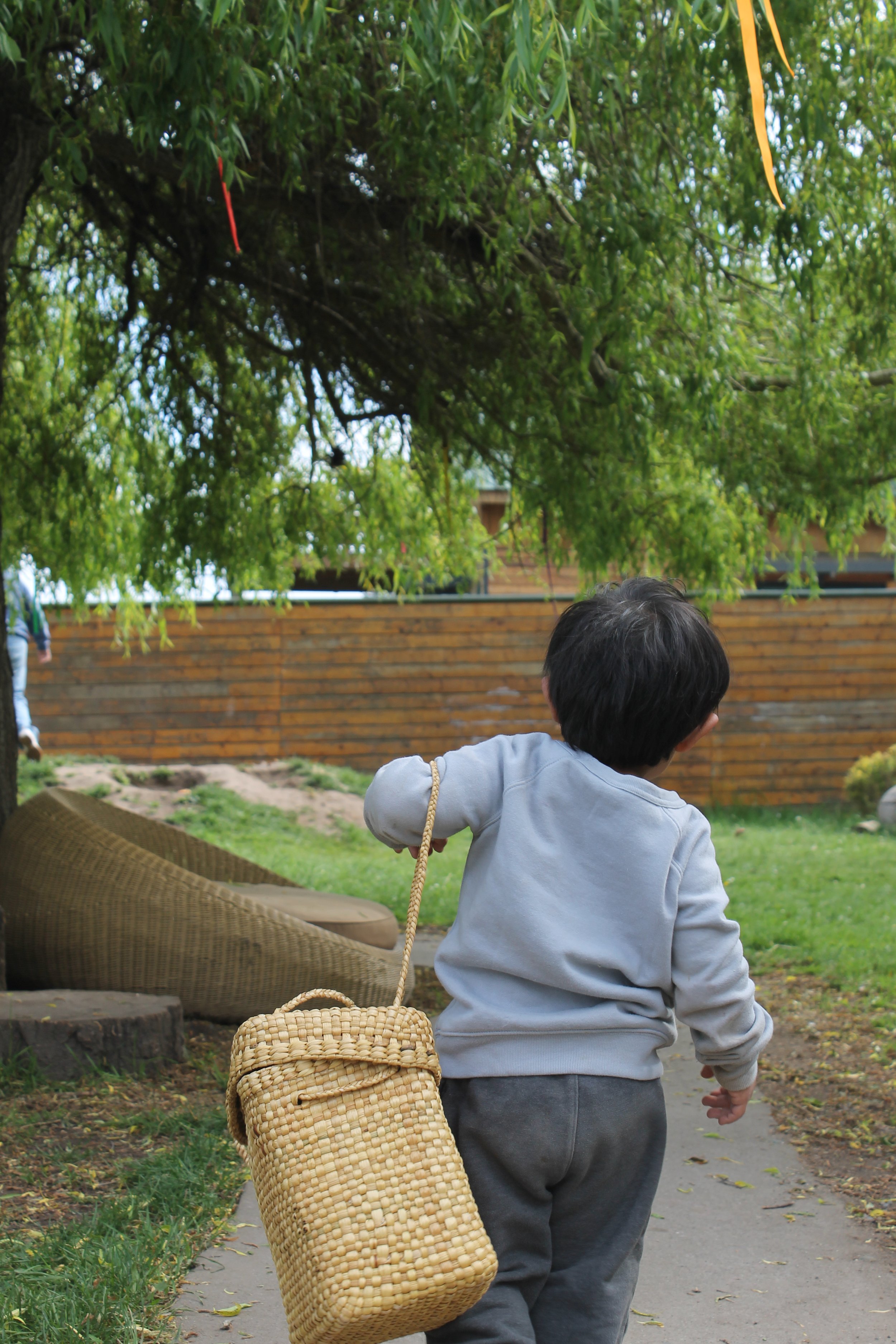 A young boy with dark hair wearing a light gray hoodie and dark gray pants, walking on a paved path in a park or garden. He is carrying a woven straw bag with a rope handle hanging over his right shoulder. The background features lush green trees, a wooden fence, and a man in the distance walking along the same path.