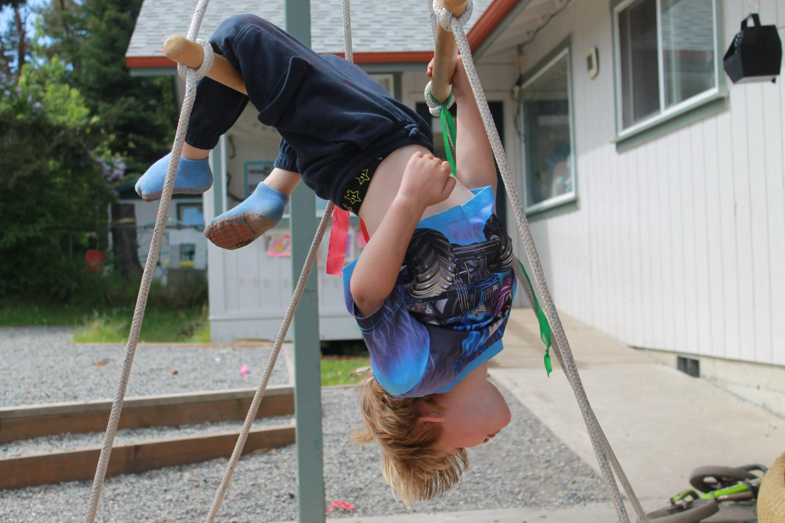 A child hanging upside down on a rope swing outdoor near a preschool with trees in the background.