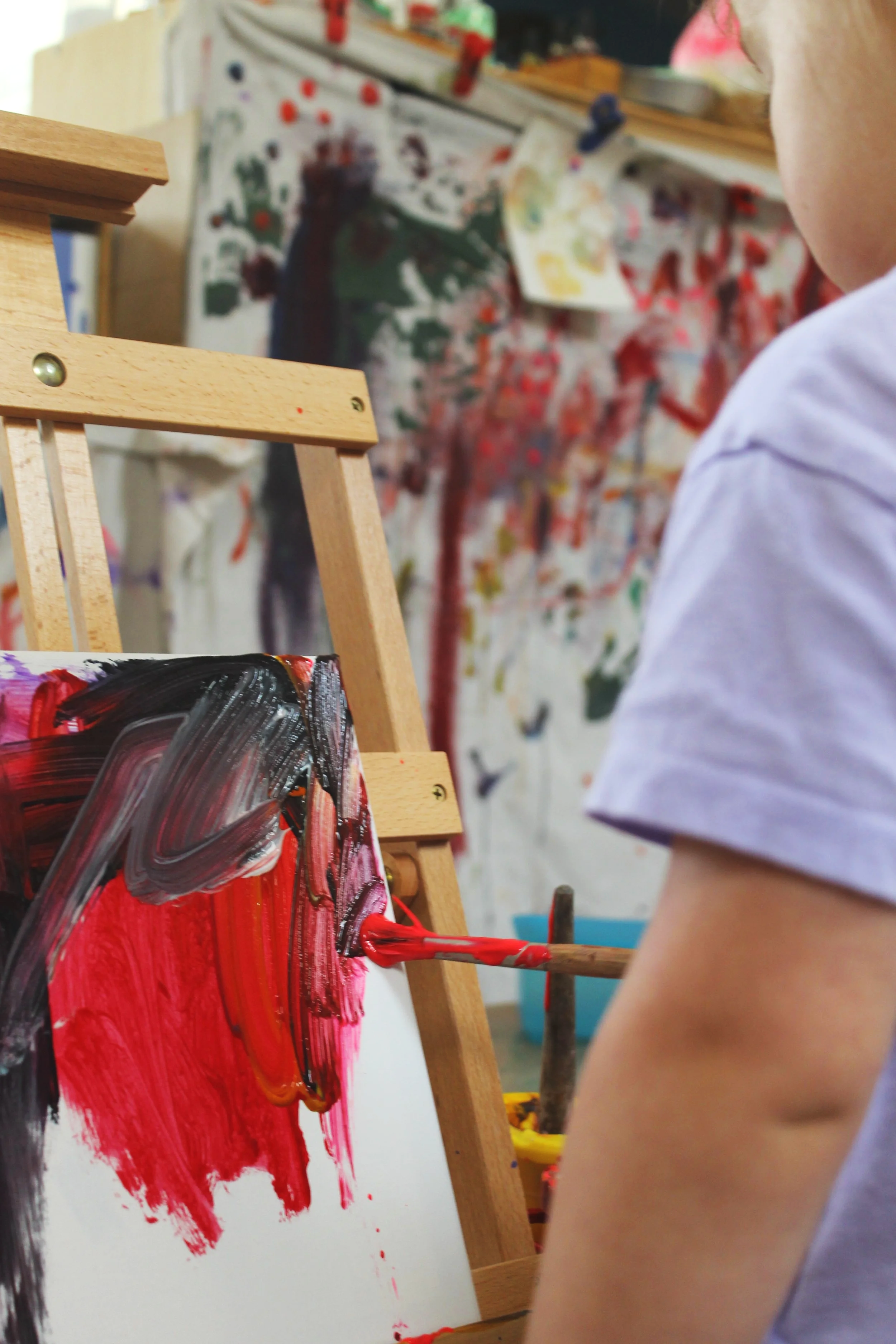 Child wearing a white shirt painting with red and black paint on a canvas, with a colorful and messy art background.