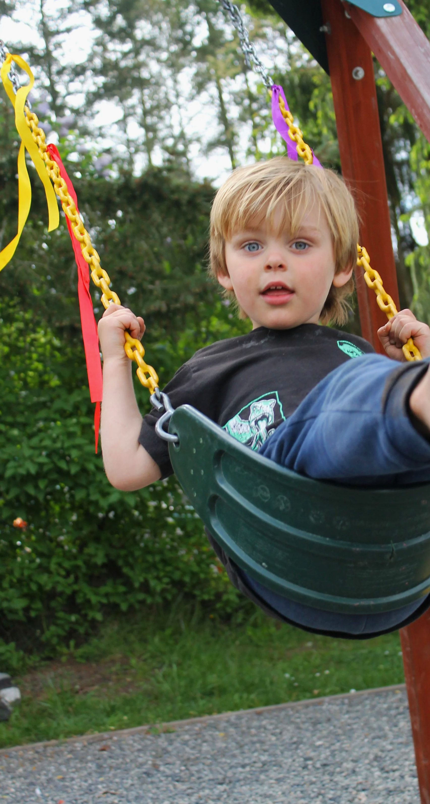 A young boy with blonde hair and blue eyes sitting on a green swing in a preschool playground, holding onto the yellow chains, with trees and greenery in the background.
