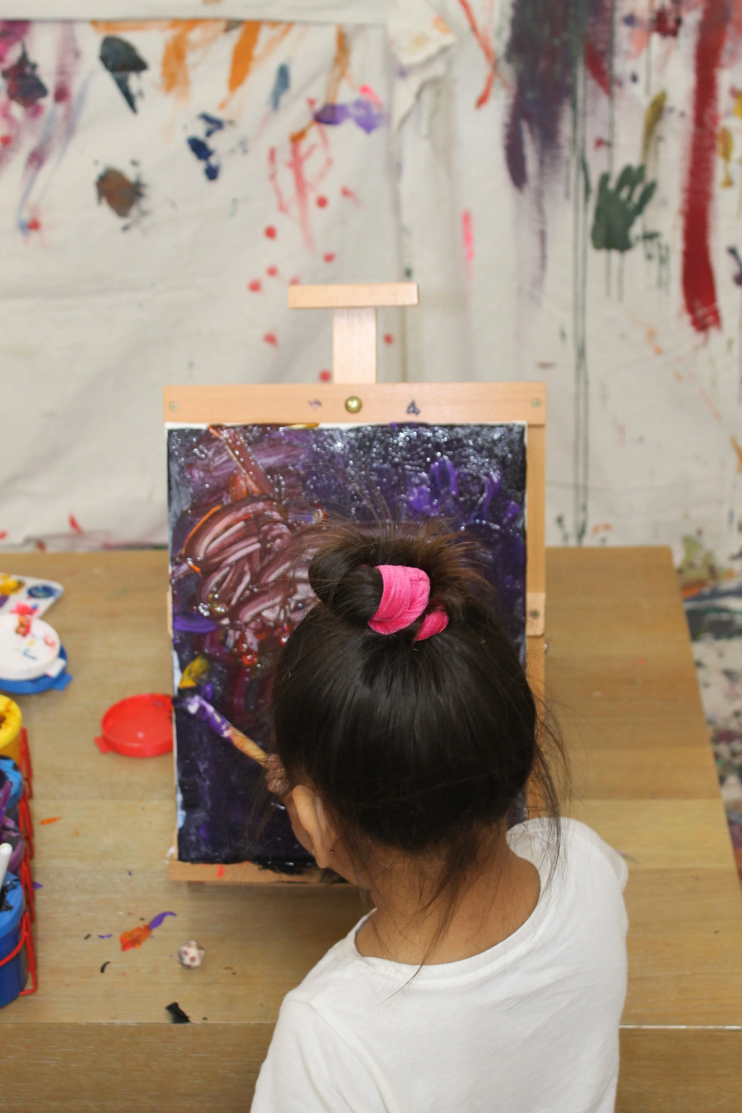 A young girl with dark brown hair tied in a pink scrunchie is painting abstract art on a canvas at a table, with various paint supplies nearby. The background features a wall with colorful, expressive paint splatters and streaks.