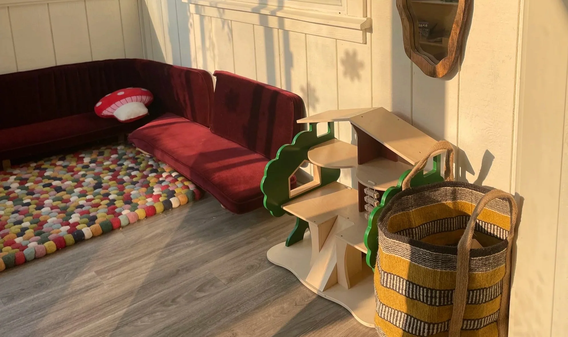 A cozy corner with a red velvet sofa, a colorful pebble-patterned rug, a wooden shelf with a green and natural wood finish, a striped woven basket, and a small wall-mounted shelf. The setting is illuminated with warm sunlight.