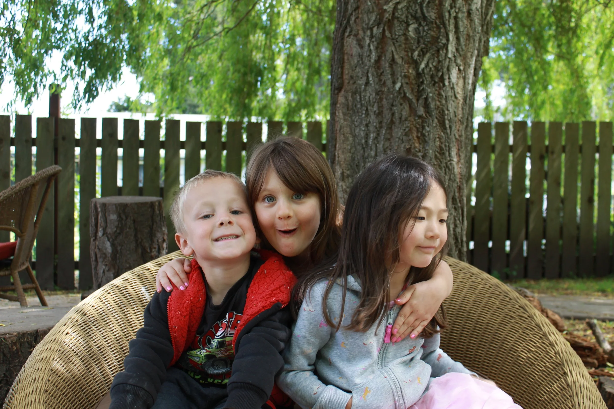 Three children sitting together outdoors on a wicker bench, with trees and a wooden fence in the background, smiling and embracing.