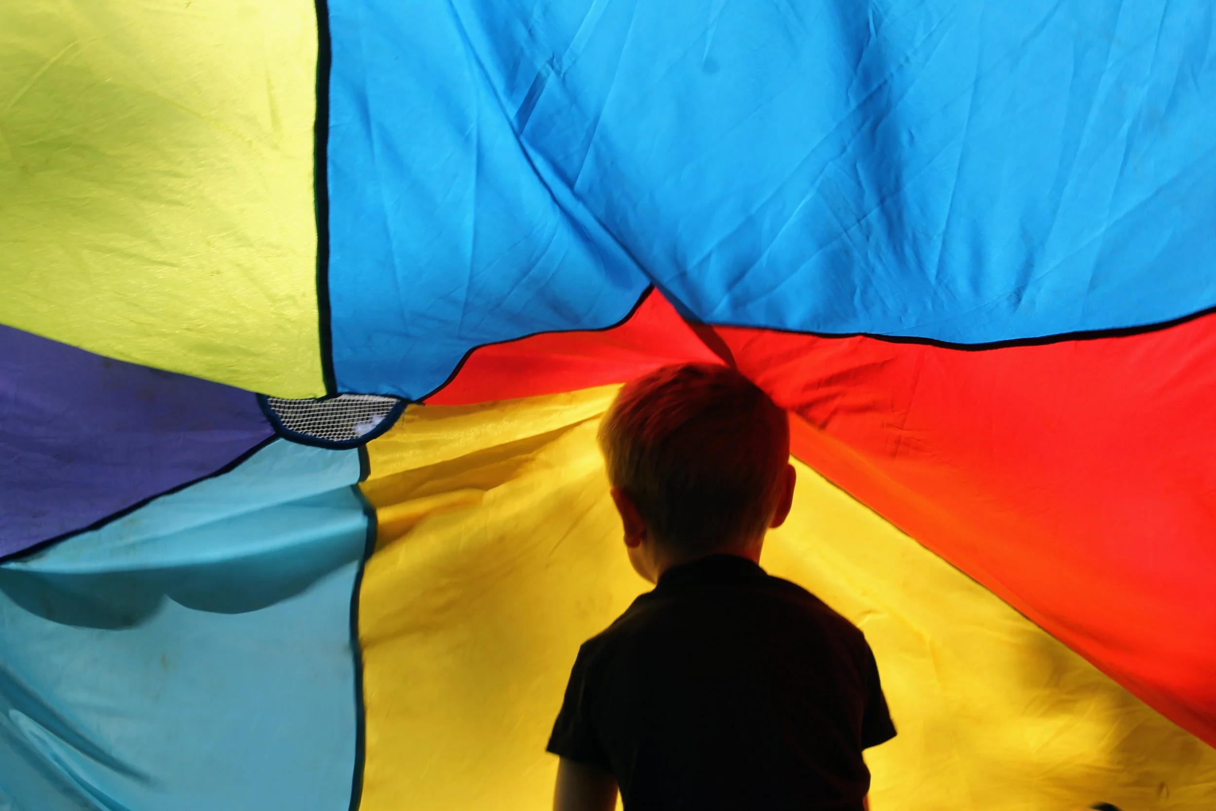 Child looking into the interior of a colorful parachute, with vibrant fabric panels in yellow, blue, red, purple, and orange.