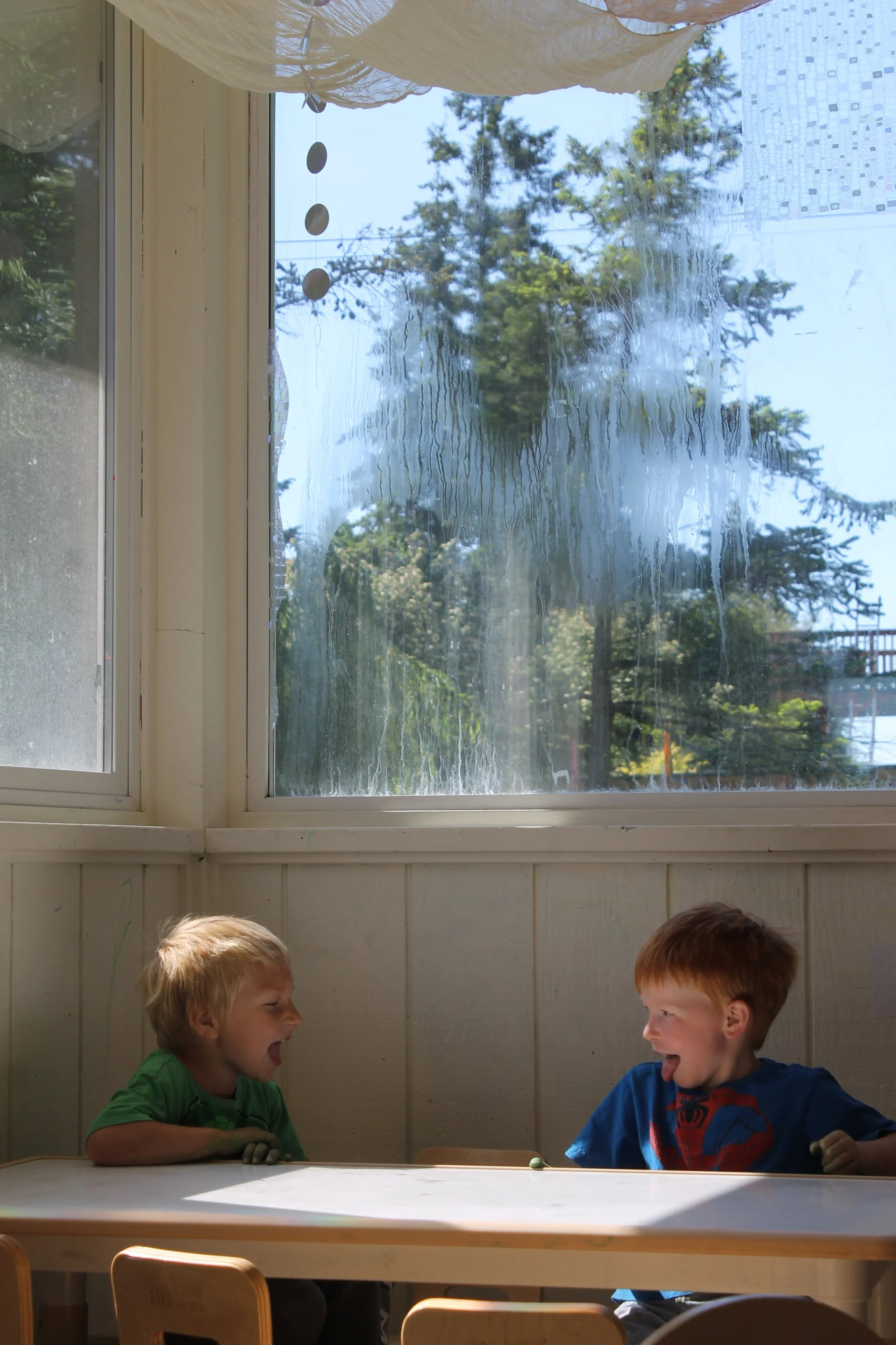 Two young boys with blonde hair sitting at a white table in a room with cream-colored walls, laughing and looking at each other. A window behind them shows a sunny day with trees outside and a cloudy sky.