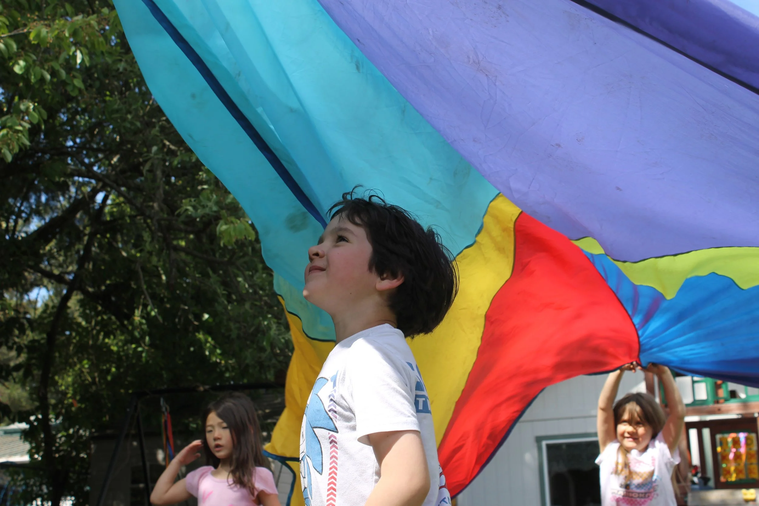 Children playing outdoors with a colorful large parachute; a young boy with dark hair and a white shirt is in the foreground, and other children are in the background.
