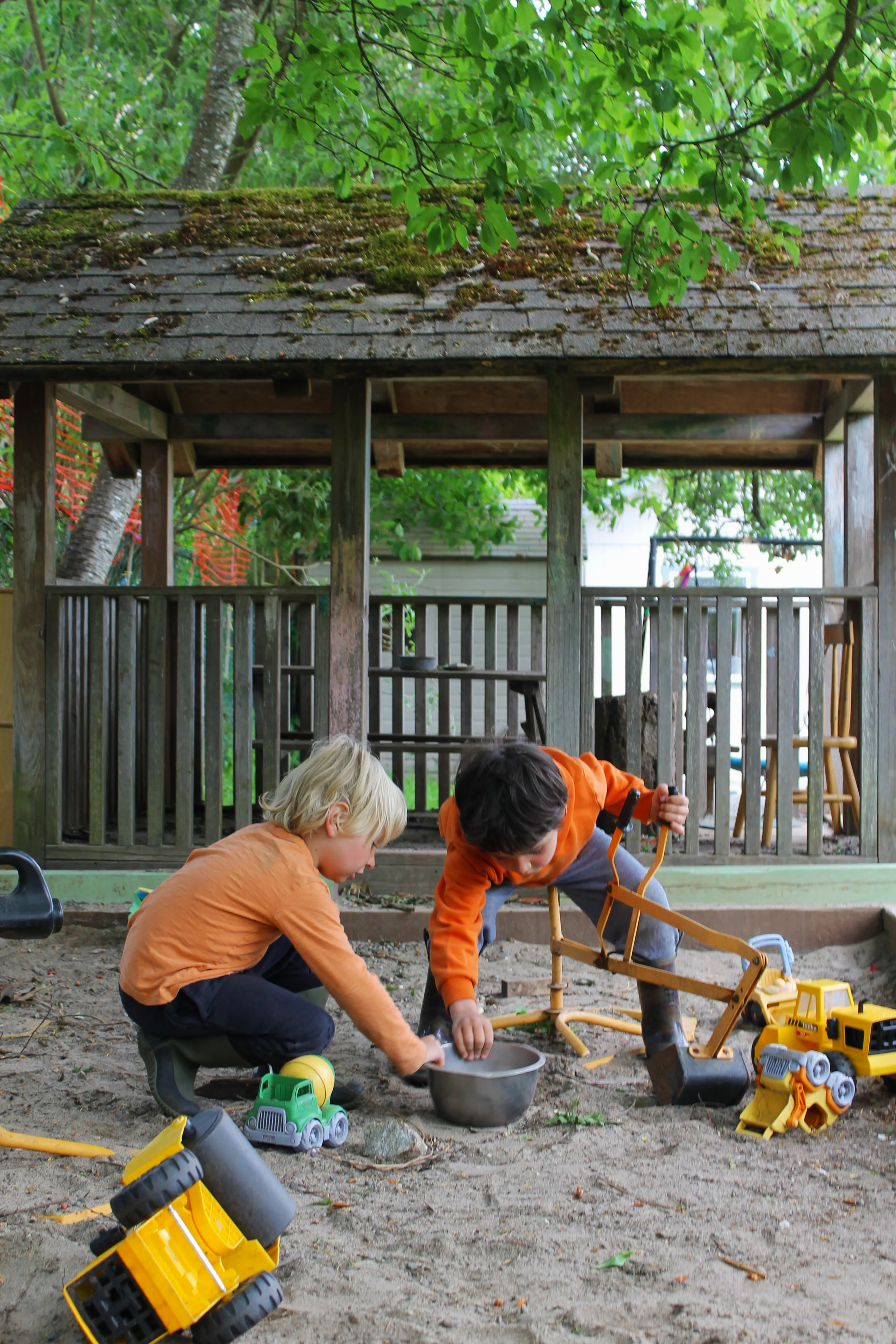Two children playing in a sandbox outdoors, digging in the dirt with toys, near a wooden playhouse and surrounded by trees.