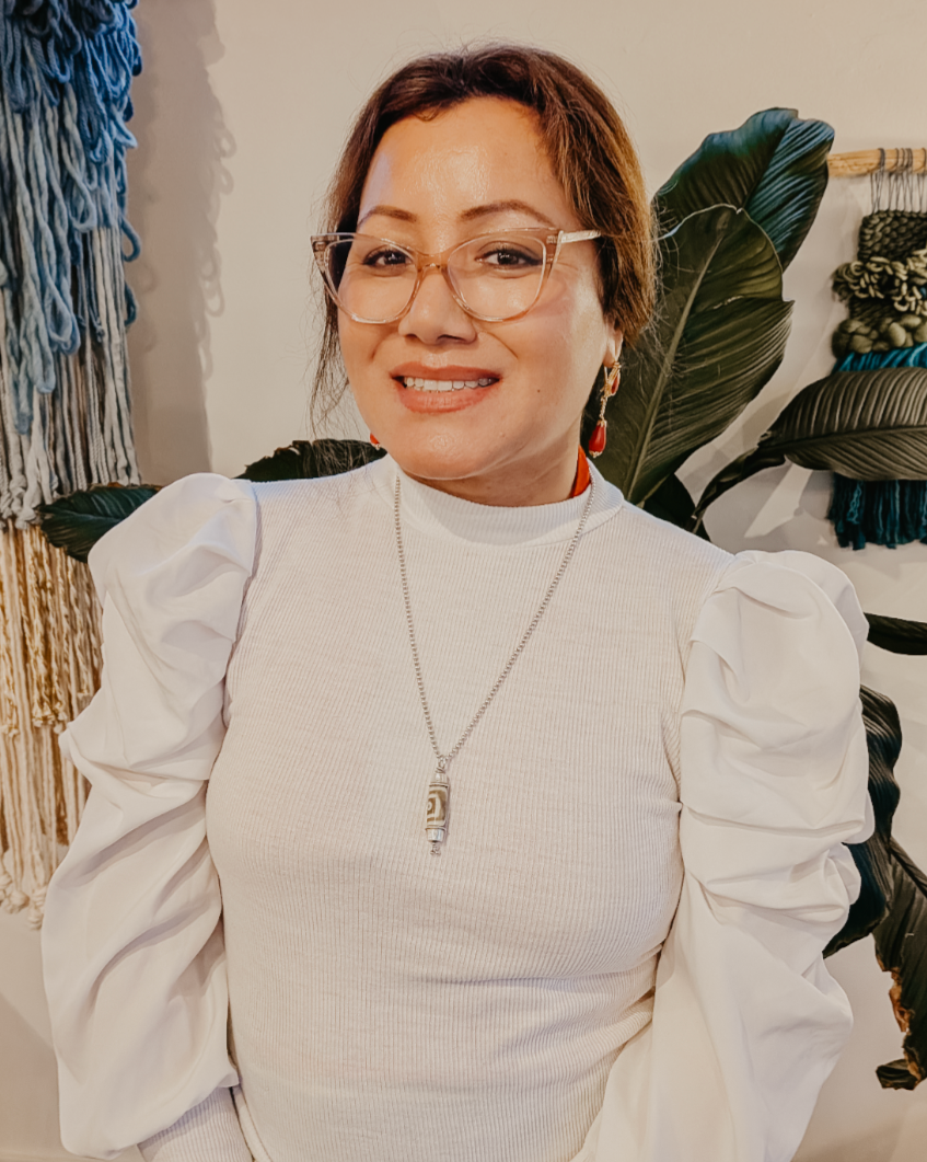 A woman with glasses on her head standing outdoors in front of a white vehicle, wearing a colorful outfit and a beige fleece jacket.