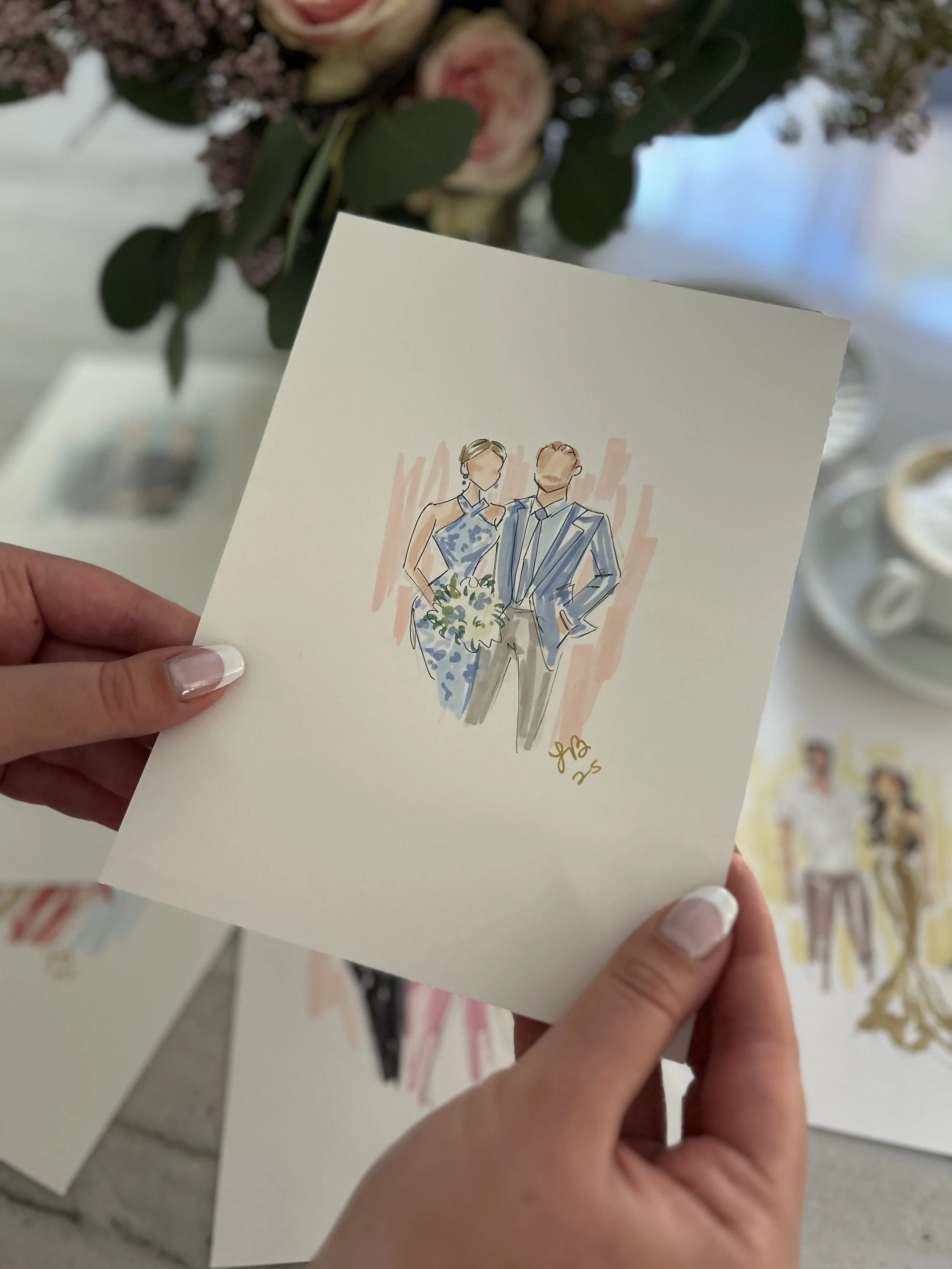 Hand holding a wedding illustration card featuring a bride and groom, with flowers in the background.