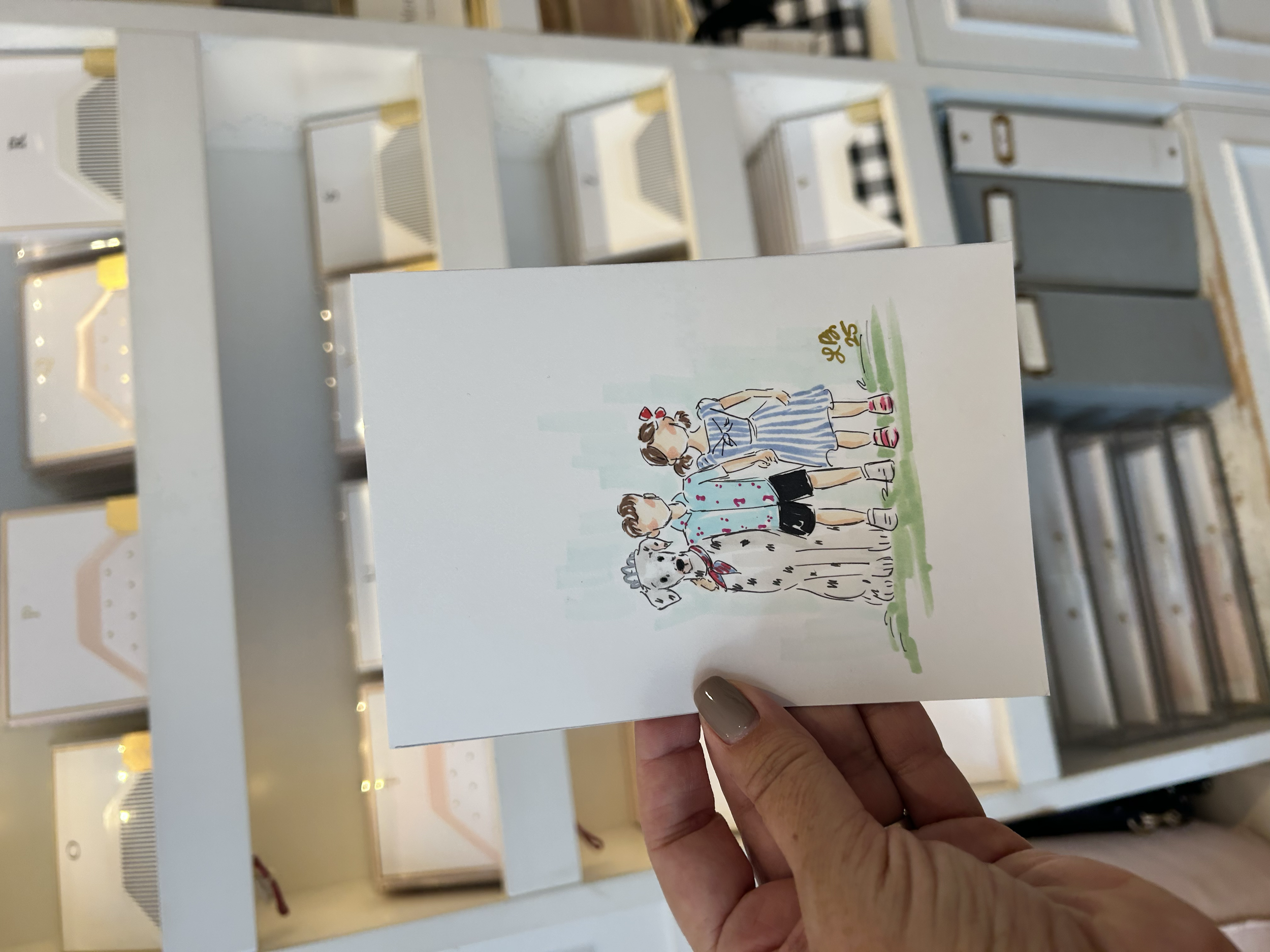 A hand holding a greeting card with an illustration of two children and a Dalmatian dog outside, with shelves of other greeting cards in the background.