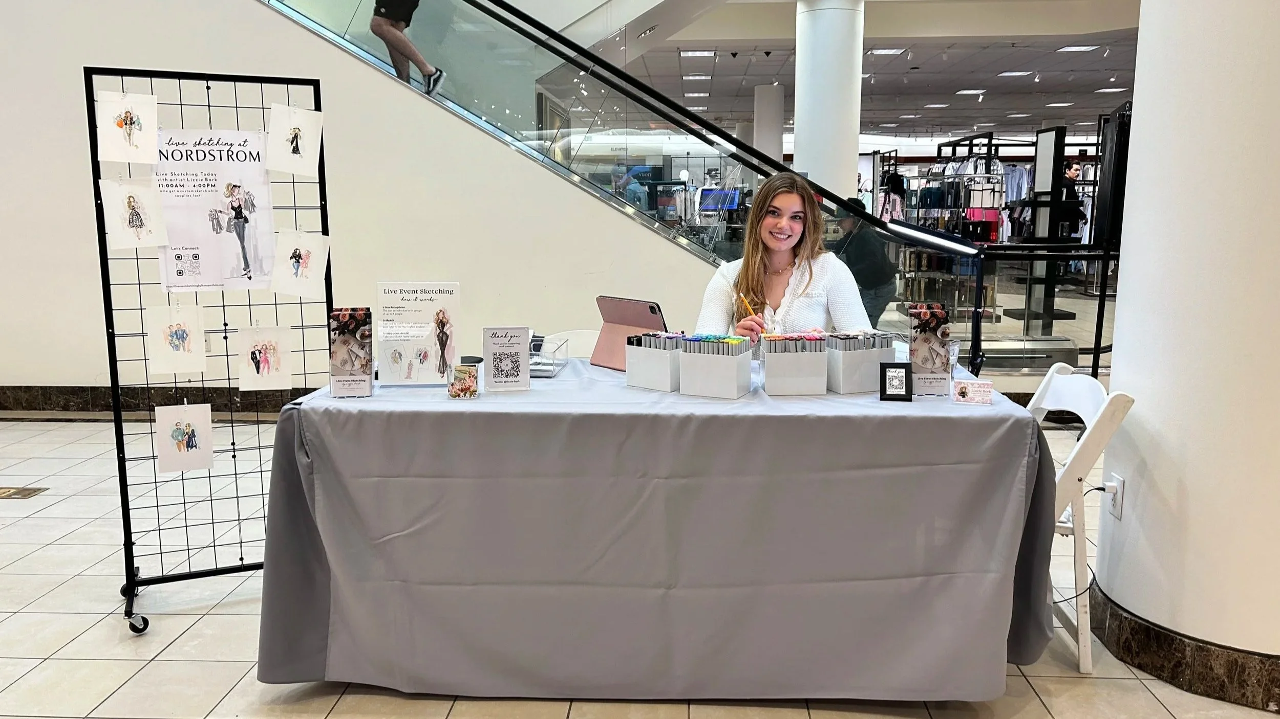 A smiling young woman sitting at a table illustrated with markers and pens inside a shopping mall, with a nylon skating event poster and a QR code on display, and a black metal grid showcasing fashion sketches to her left.