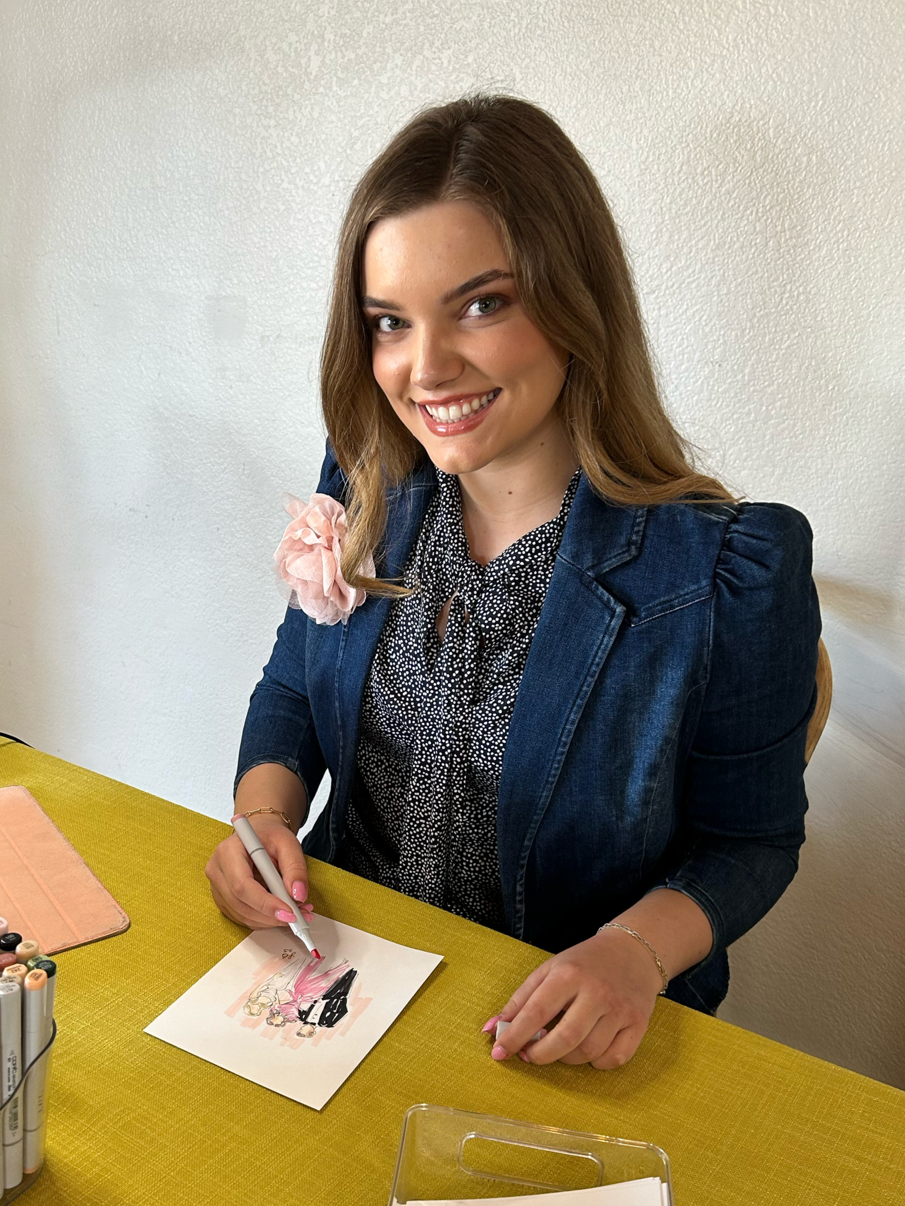 A young woman sitting at a table, smiling and drawing or signing on a piece of paper with markers, wearing a denim jacket and a polka dot blouse.