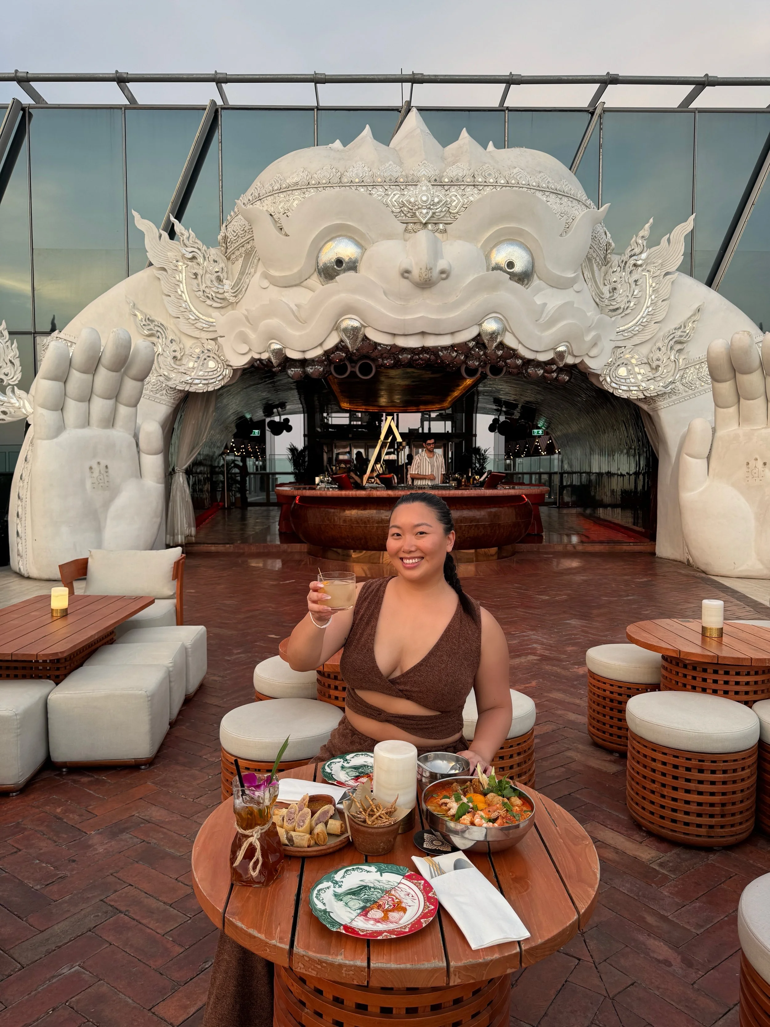 A woman smiling and holding a drink, seated at a round table with food including sushi, a salad, and drinks, in an outdoor setting with a large white decorative dragon arch and a DJ in the background.