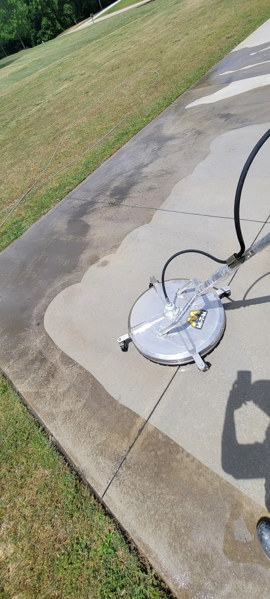 A sidewalk power washer cleaning concrete, with wet and dry areas visible, next to a grassy yard with trees in the background.