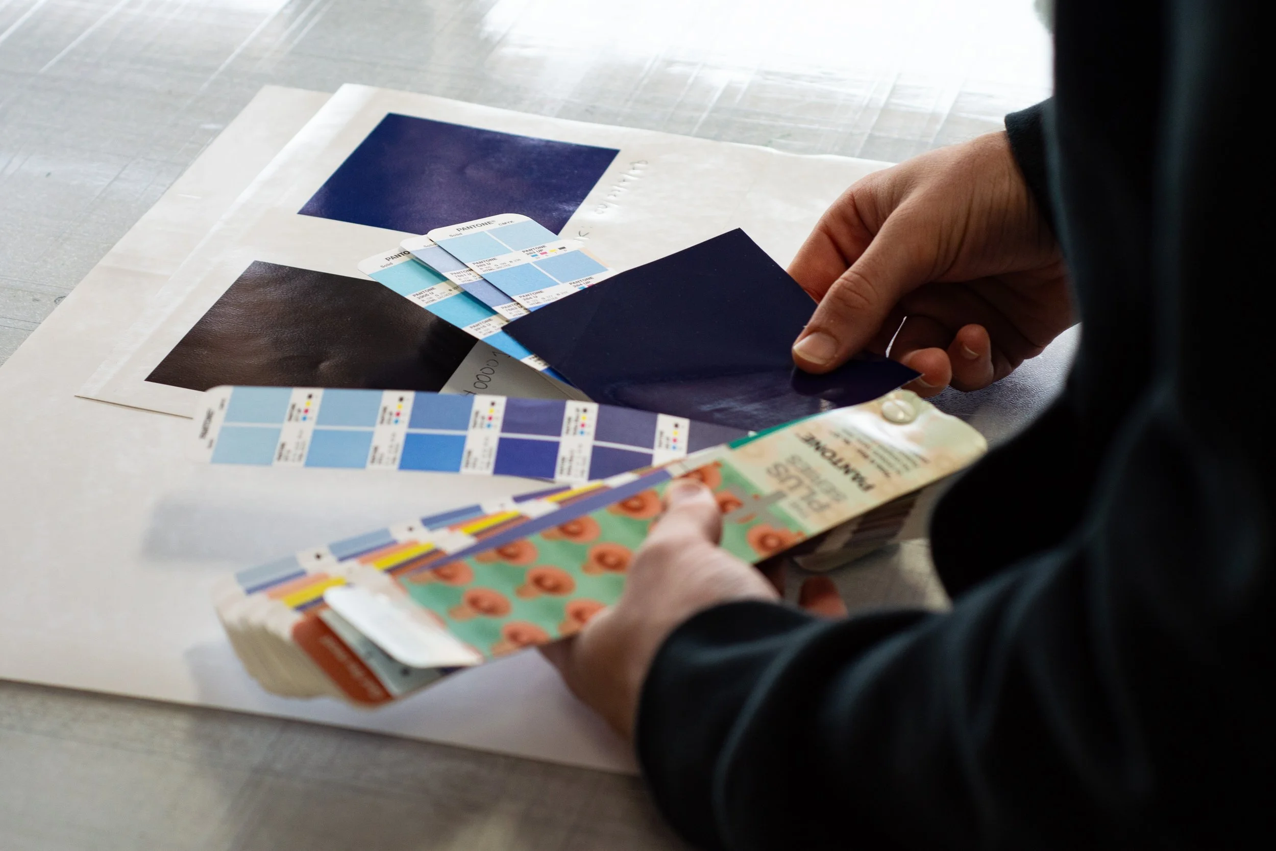 Person holding a color swatch and paint sample cards, with sample color sheets and printed materials laid out on a white surface.