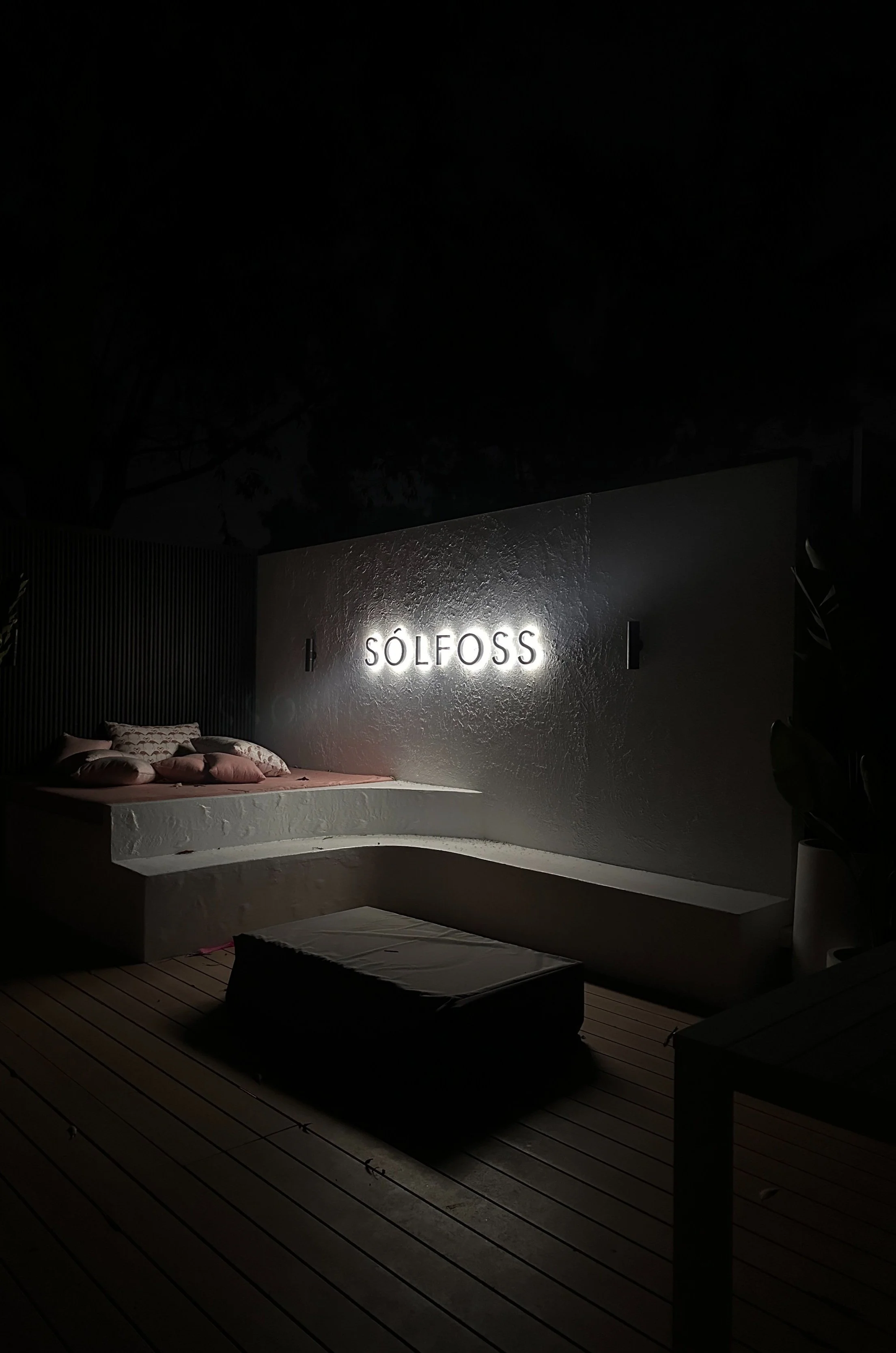 Nighttime outdoor patio with illuminated sign reading 'SÖLFOSS' on a textured white wall, a built-in corner seating area with cushions, a small table or ottoman, and a wooden deck floor.