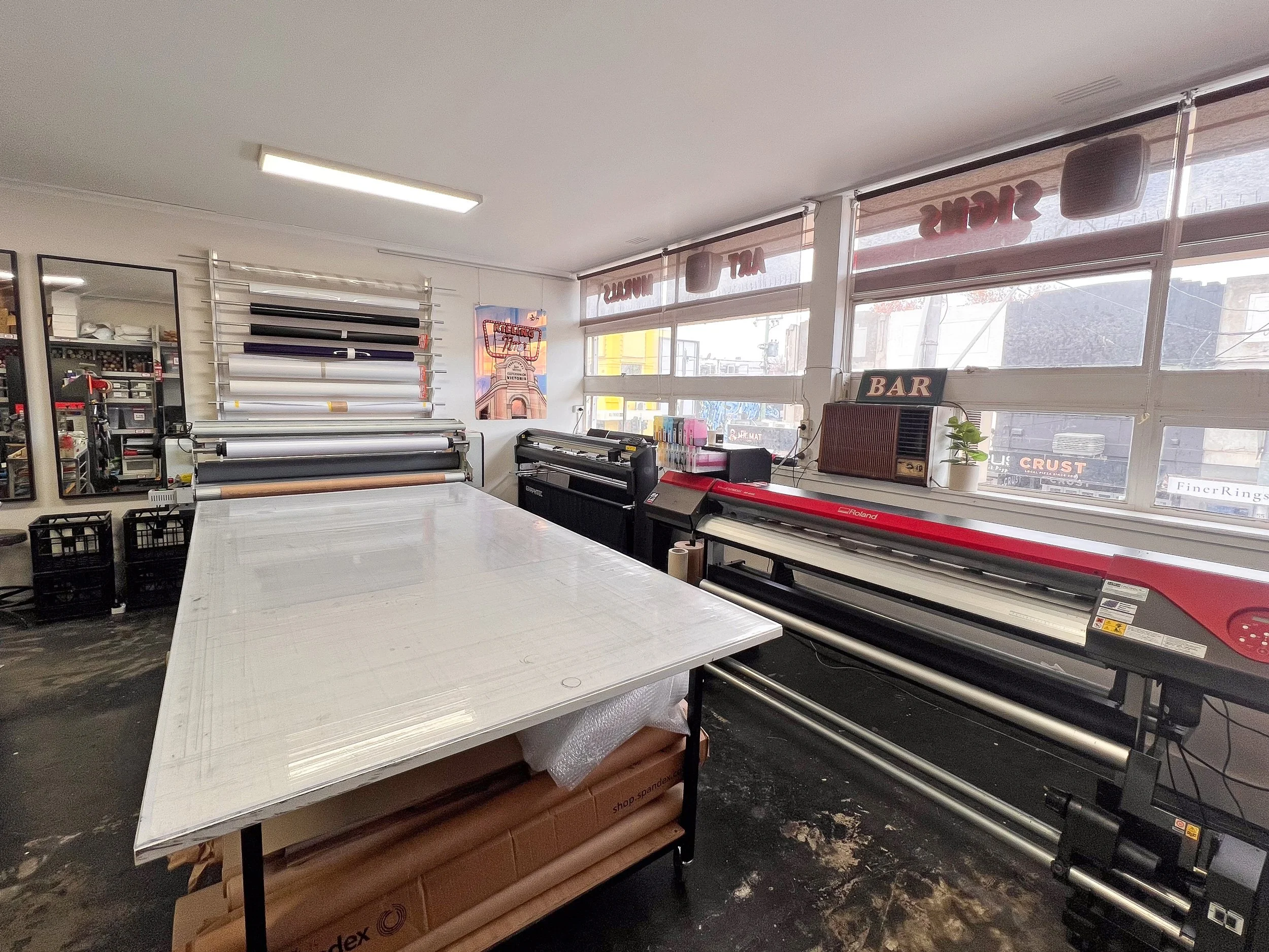 Interior of a sign shop with large cutting table, printers, and rolls of vinyl in front of windows.