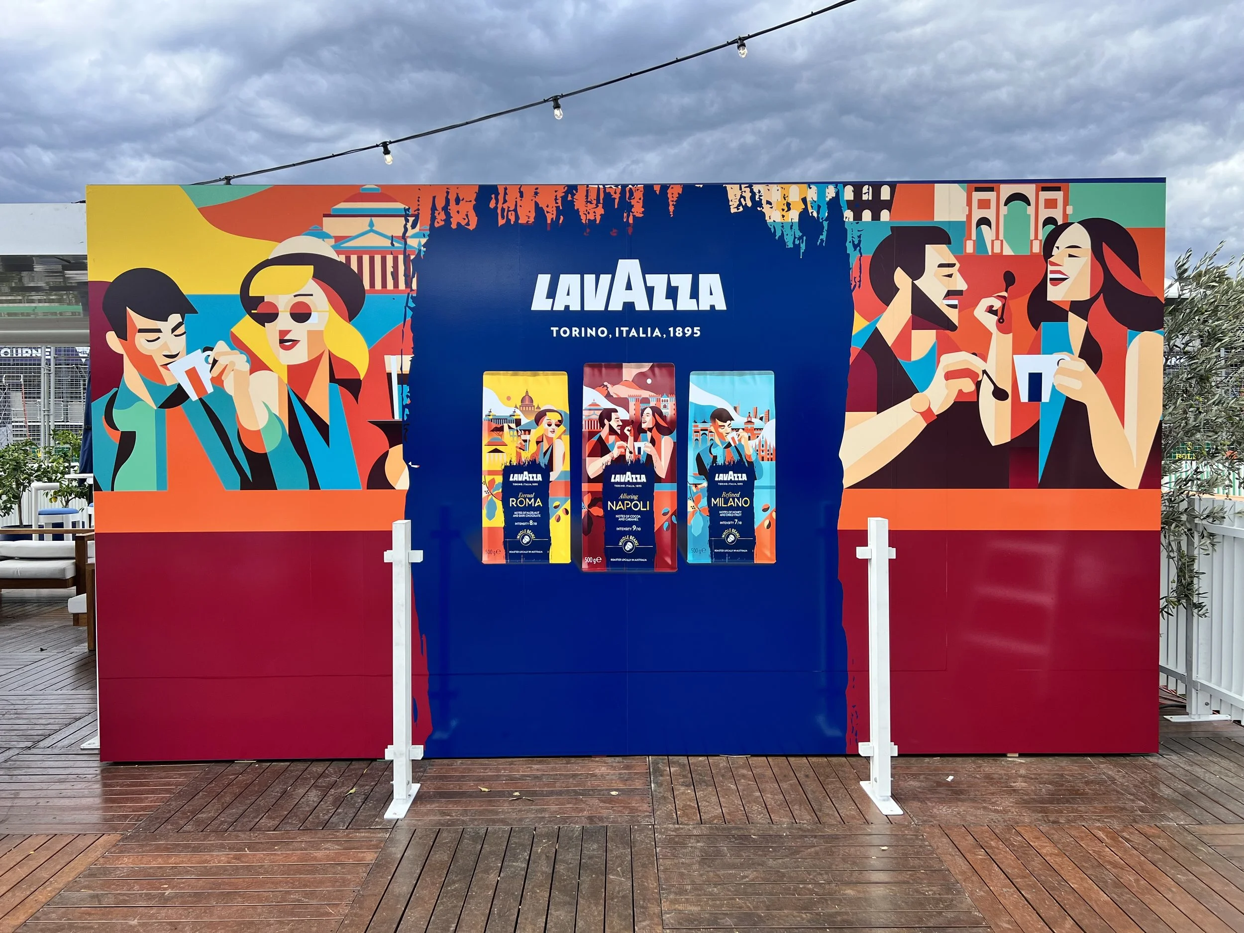Colorful advertising wall for LavAzza coffee featuring illustrations of people drinking coffee, with the LavAzza logo and the words 'Torino, Italia, 1895' in the center. There are three coffee bag images showing different blends.