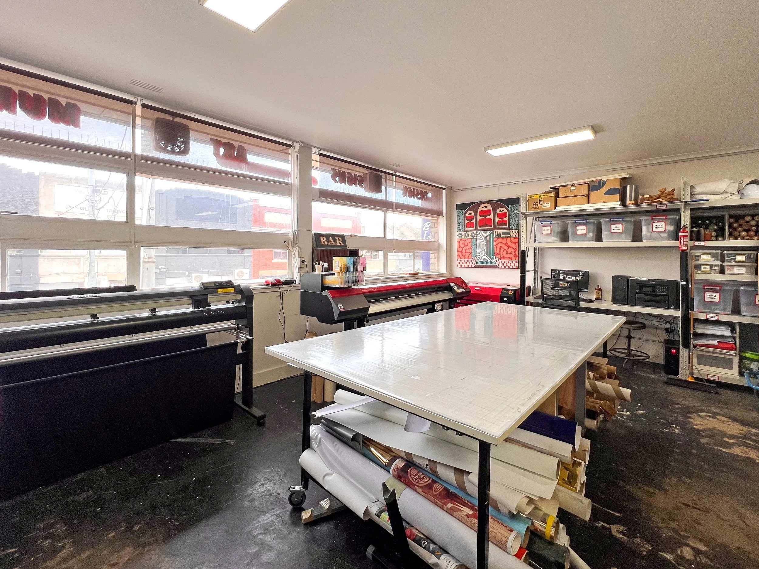Interior of a creative workspace or art studio with printing equipment, shelves with storage bins, and a table with design plans.