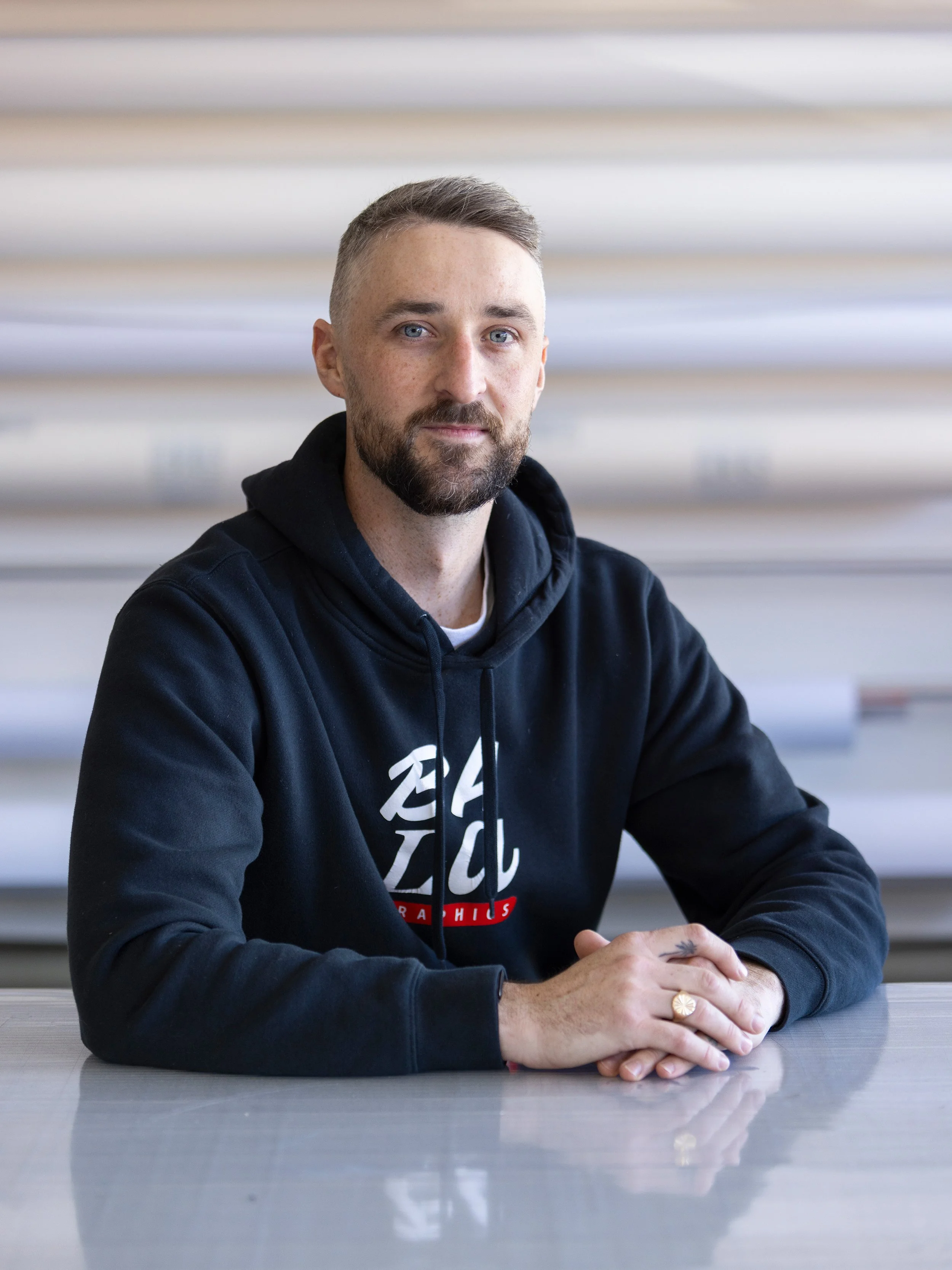 A man with a beard and short hair, wearing a black hoodie with a logo, sitting at a table in an indoor setting with blurred background.
