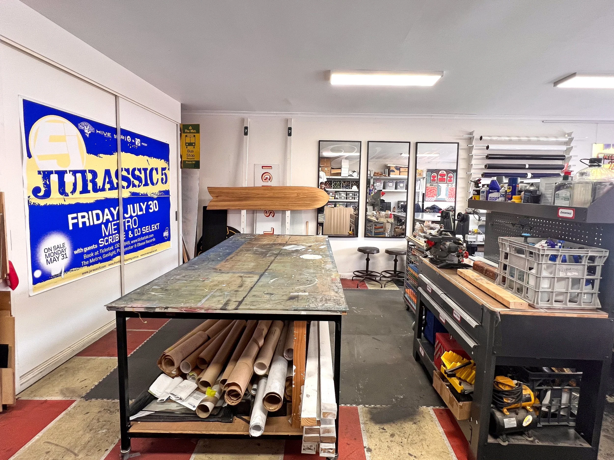 Workshop with workbenches, tools, and storage shelves, including a large calculator, rolls of paper, and a wall poster for a Jurassic concert event.